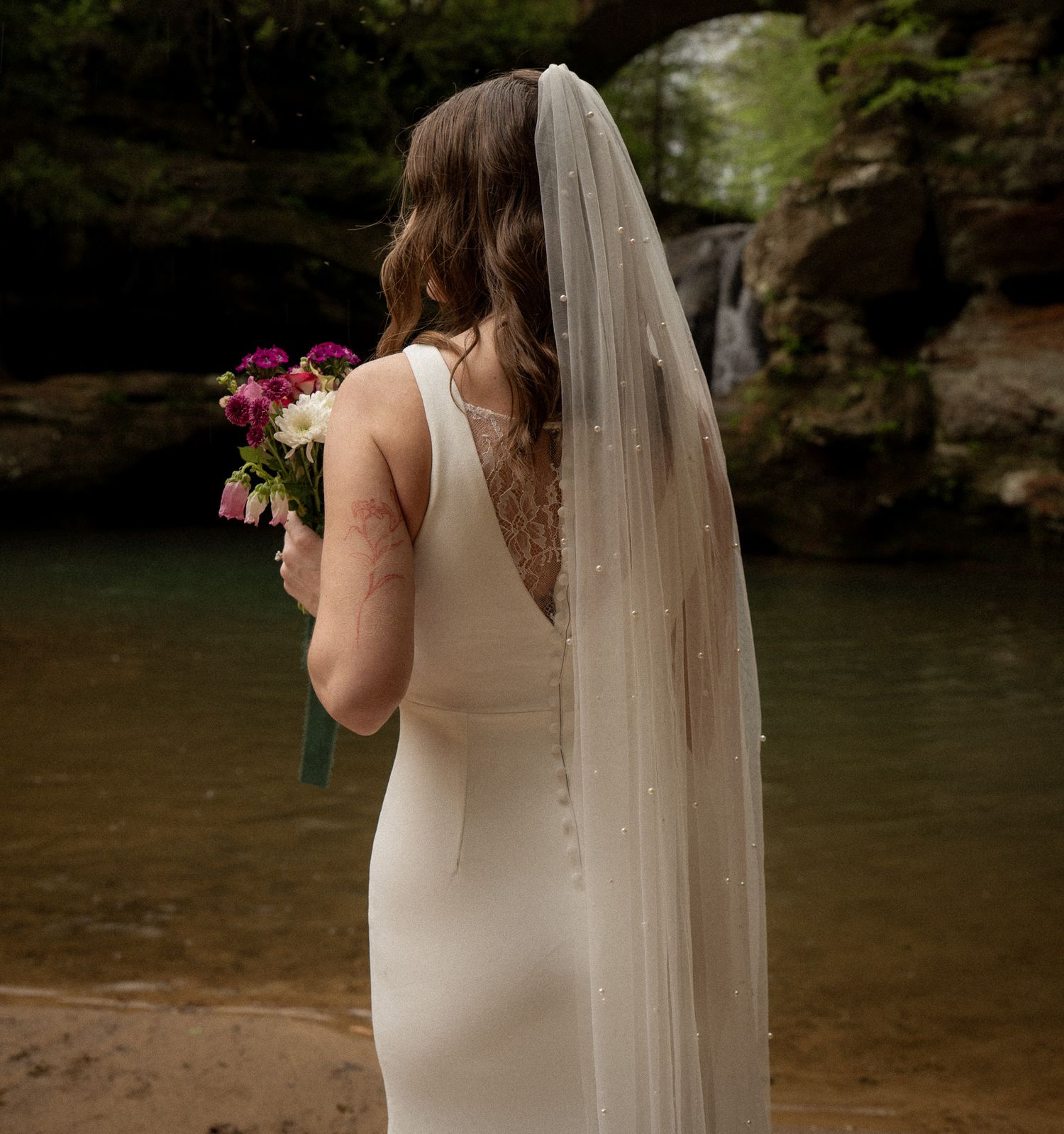 Bride in a sleek gown and veil holding a vibrant bouquet, standing by a tranquil river with a waterfall in the background.