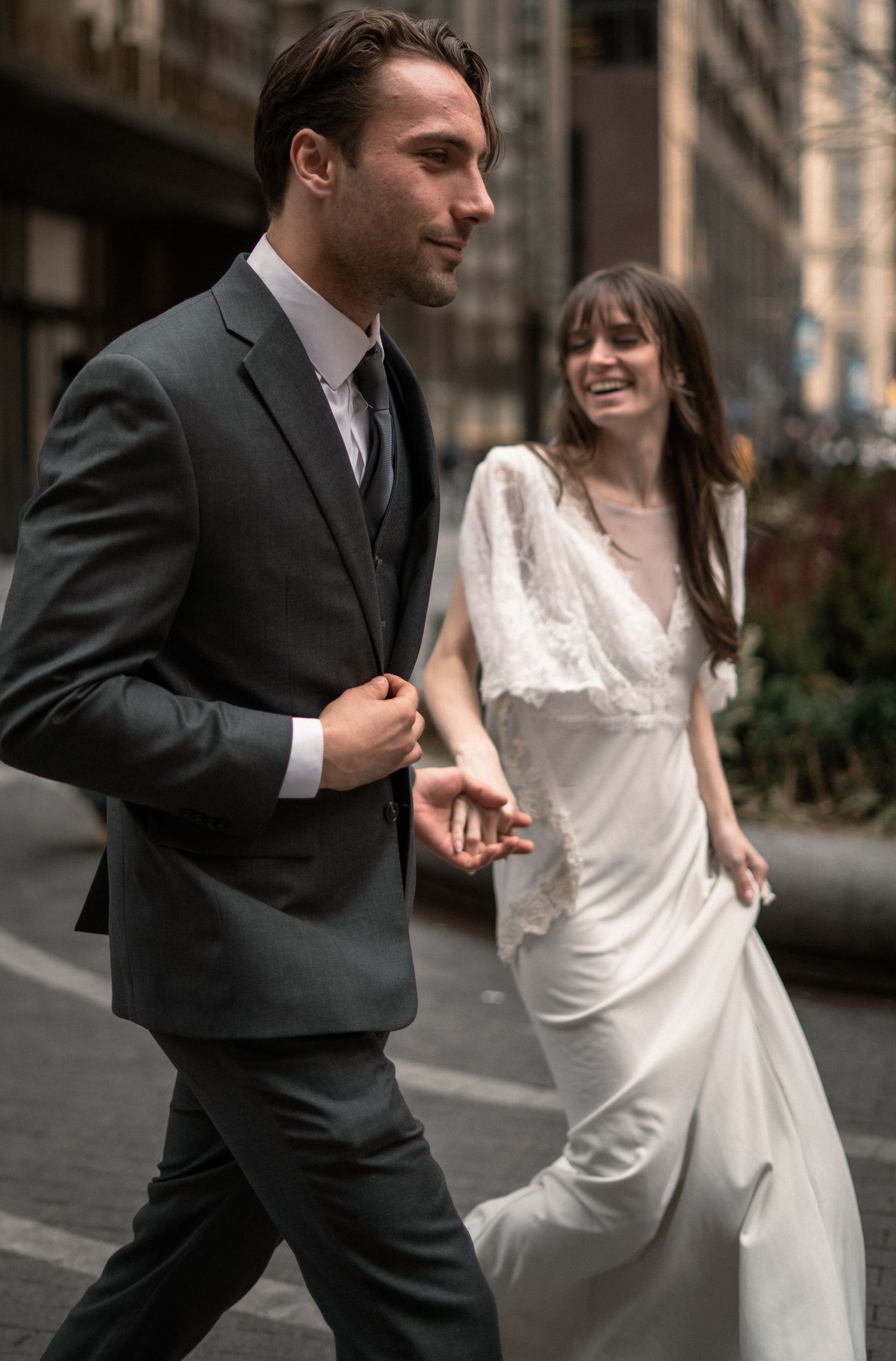 Couple in wedding attire walking together on city sidewalk with tall buildings in background.