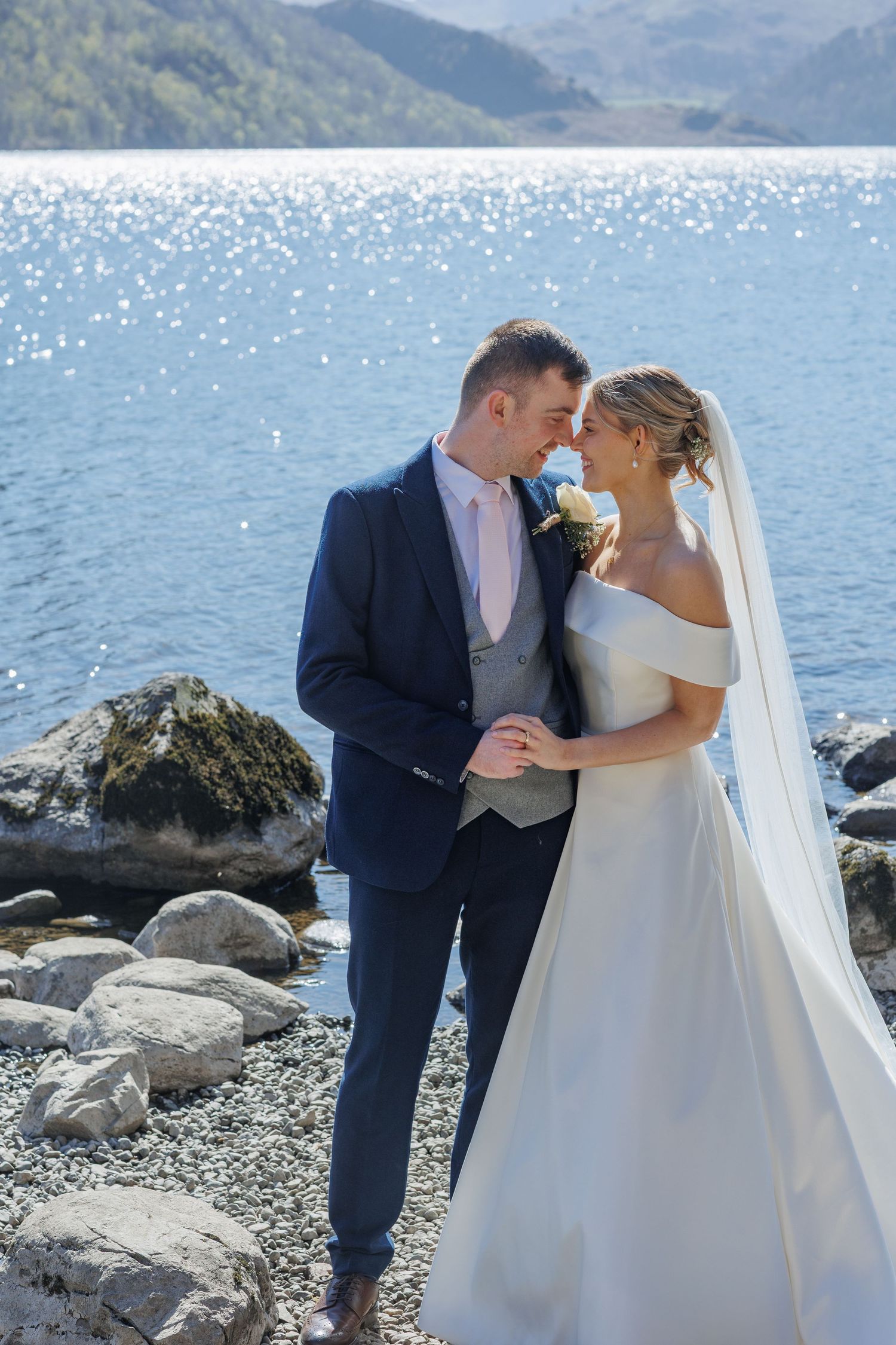 A couple shares an intimate moment by a sparkling lake with mountains in the background.