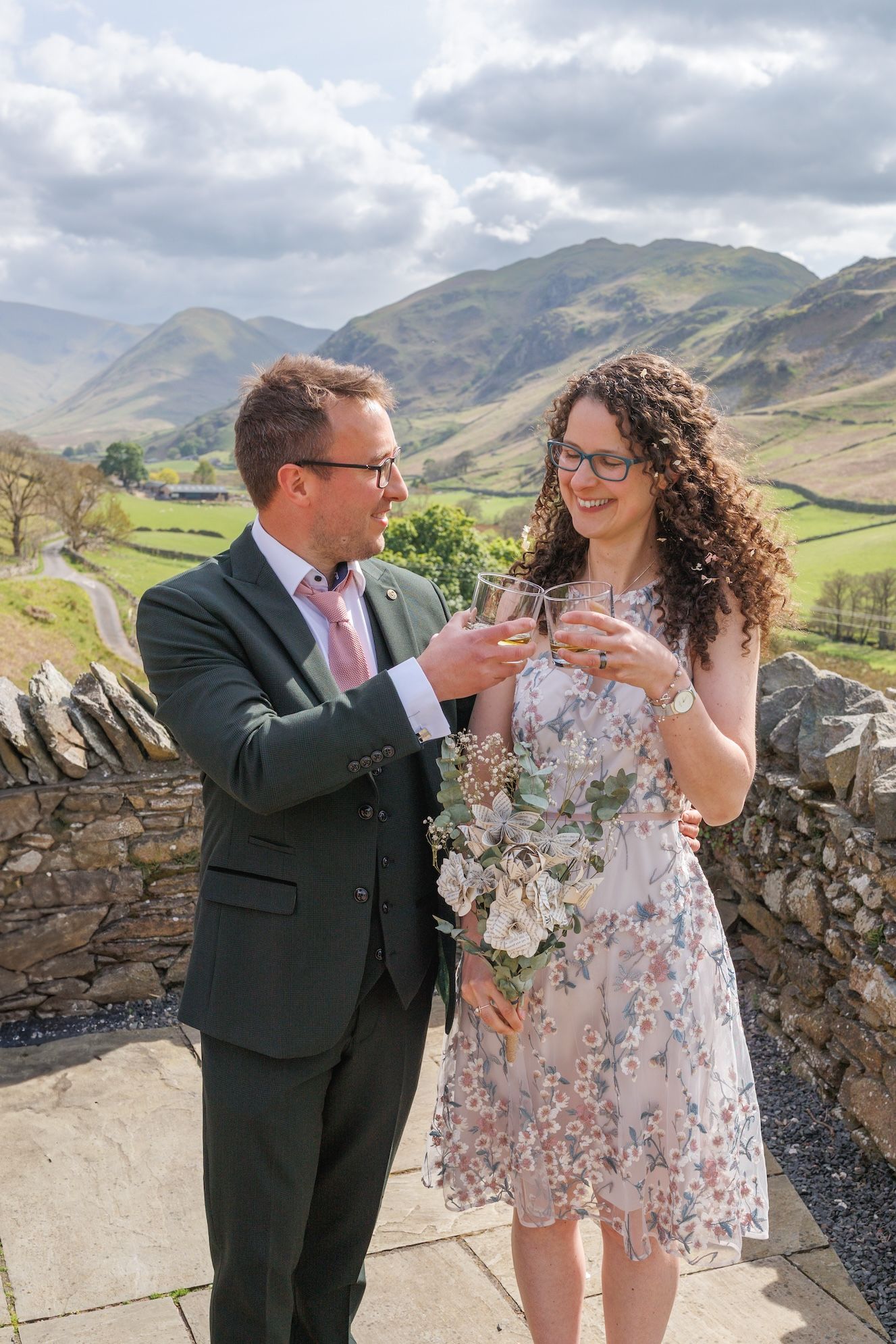 Two people toast with drinks against a scenic Lake District mountain backdrop.