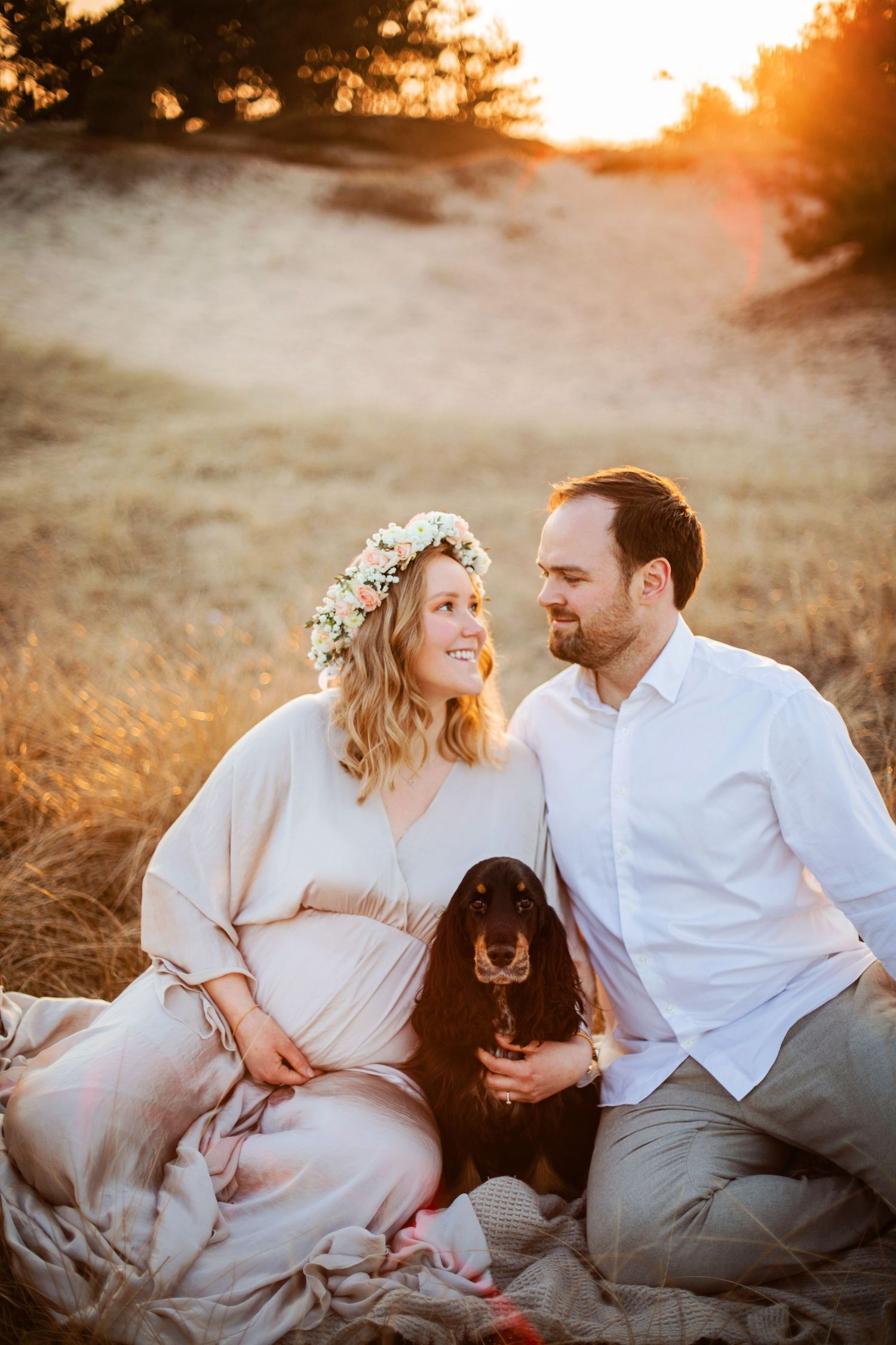 A couple sits on a blanket with their black dog during sunset in a golden field while wearing light colored clothing.