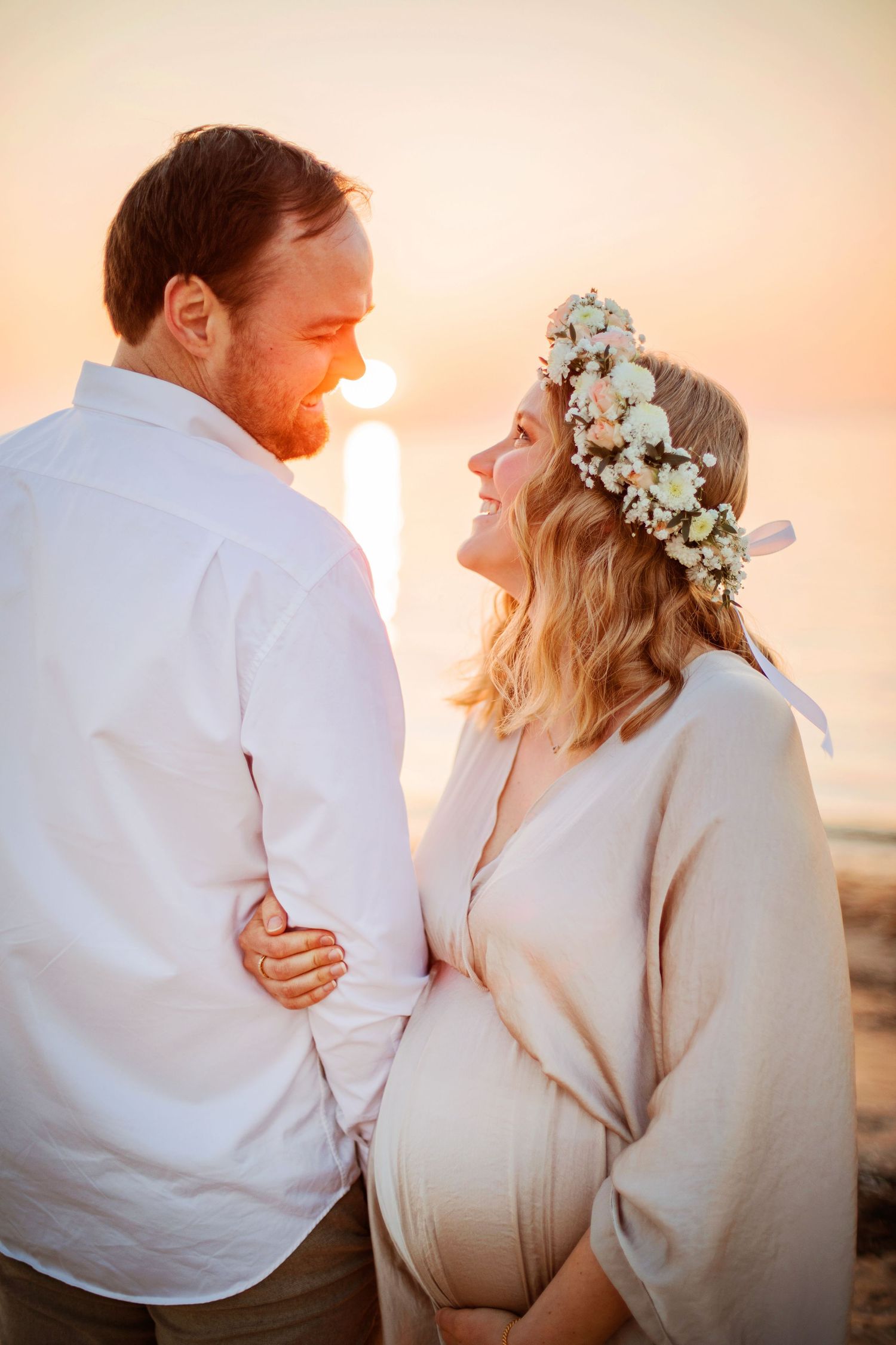 Romantic outdoor sunset photoshoot showing expectant couple in white attire with woman wearing floral crown looking at each other.