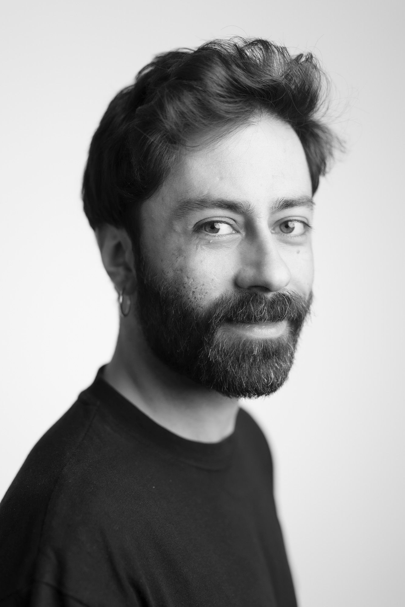 Black and white studio portrait showing a smiling bearded person wearing a dark t-shirt against a light background.
