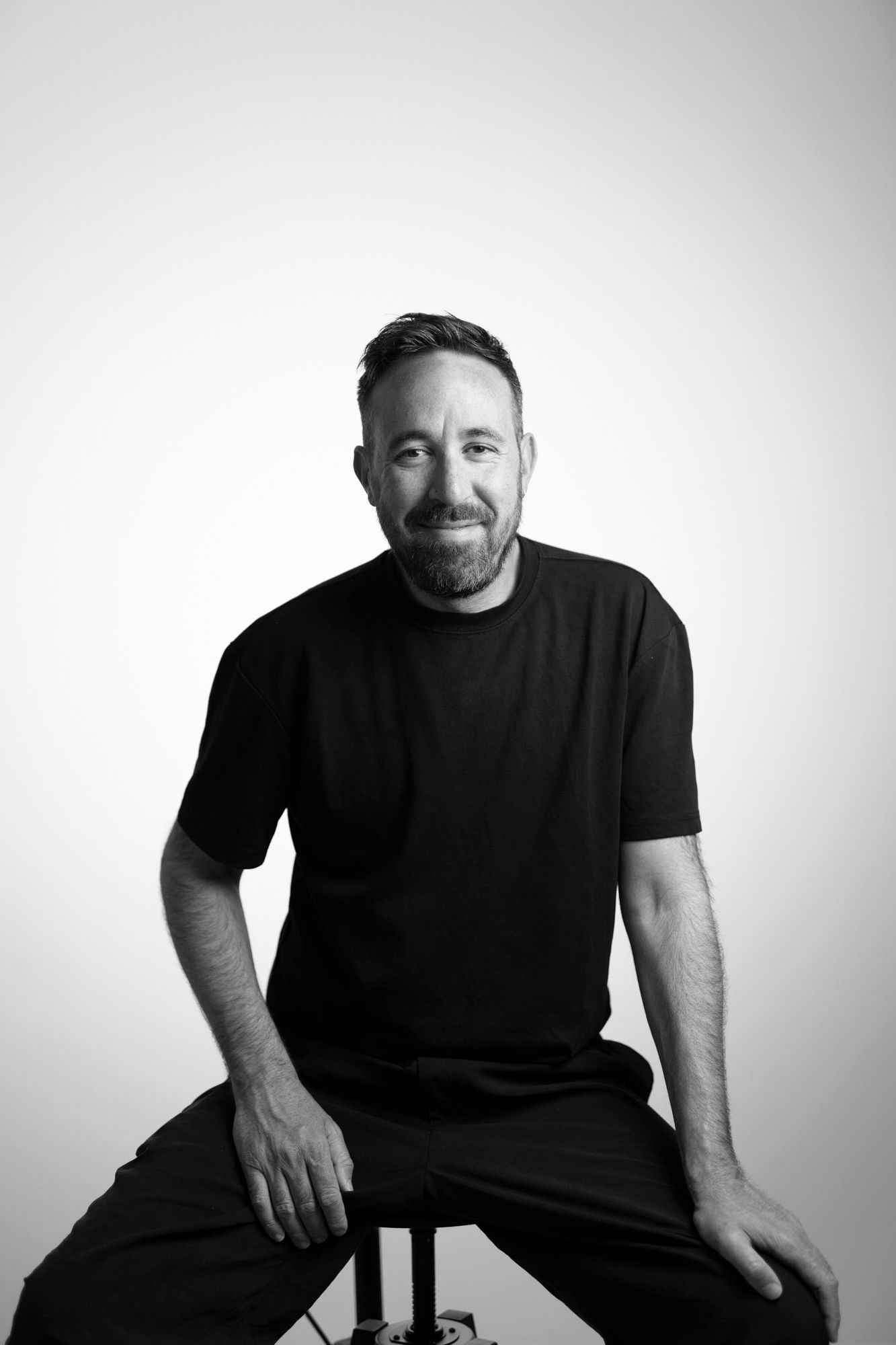 Black and white studio portrait of a bearded person in dark clothing seated on a stool against a light background.