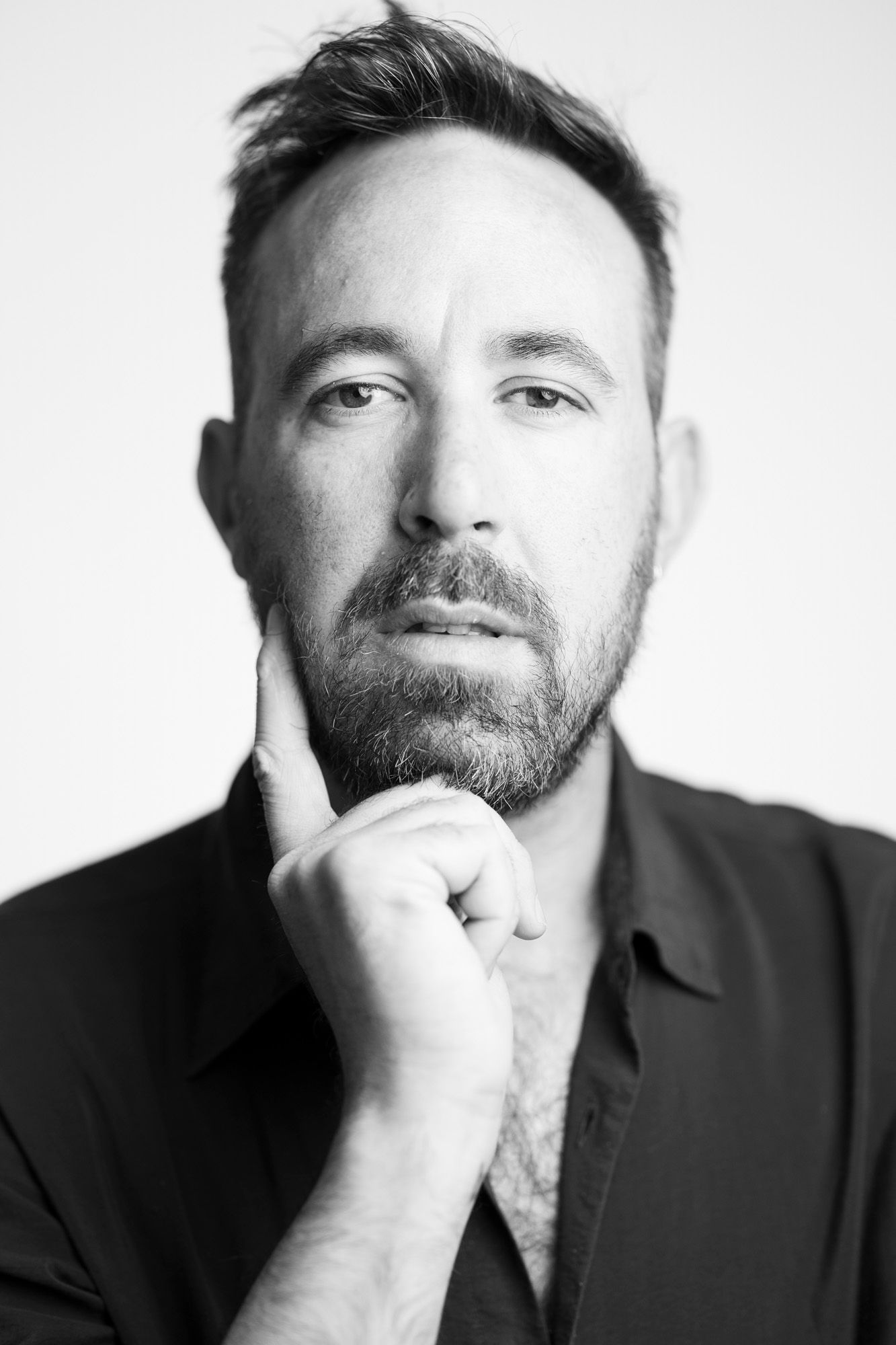 A black and white portrait photograph shows a bearded man in dark clothing with hand thoughtfully placed on chin.