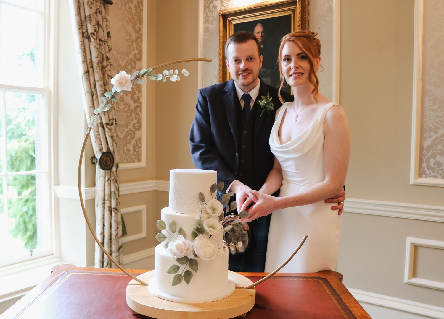 A couple cuts their elegant white wedding cake in a bright sunlit reception room while guests look on.
