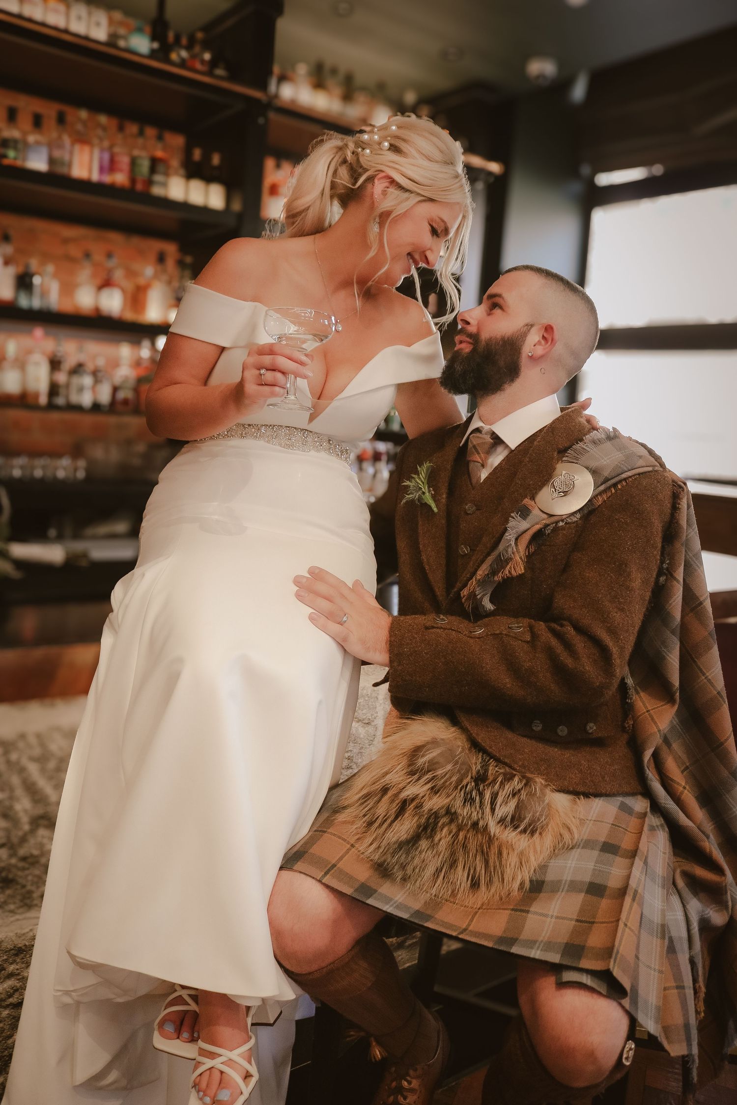 A bride in white dress and groom in traditional Scottish kilt share a romantic moment at a bar with whiskey bottles behind them.