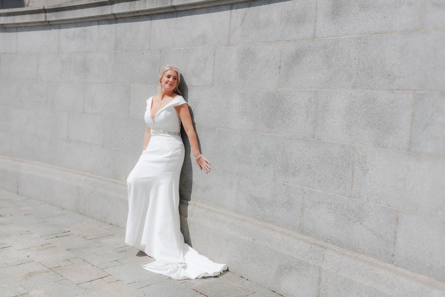 A woman in an elegant white wedding dress leans against a gray concrete wall with a flowing train trailing behind her.