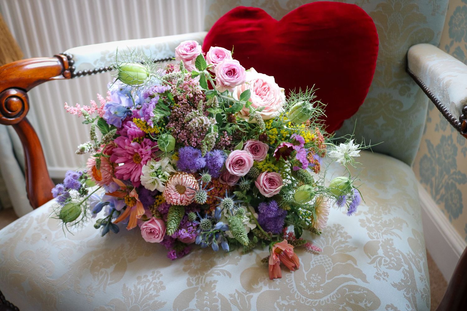 Beautiful floral arrangement with pink roses, purple flowers and greenery sits on a cream chair with red heart pillow.