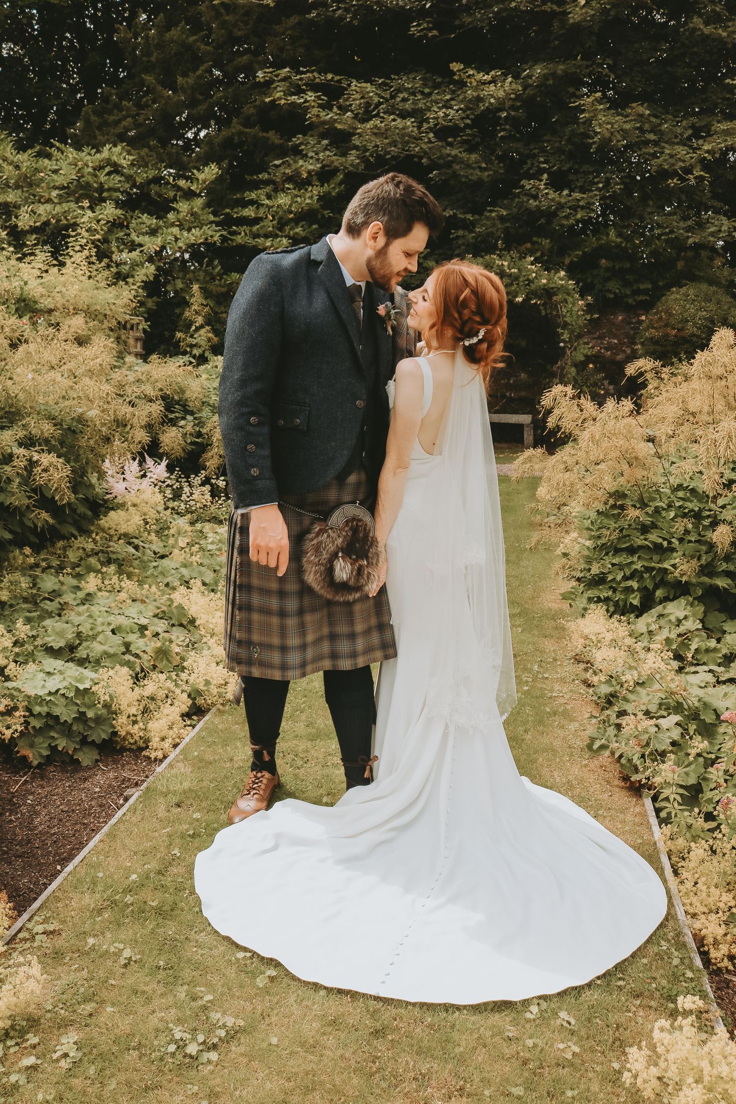 A couple in wedding attire stands together in a lush garden setting with dramatic white wedding dress trailing behind.
