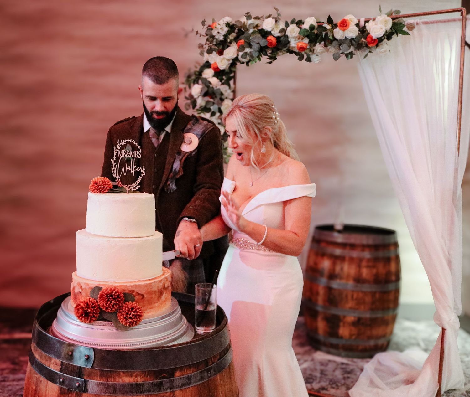 A couple cuts their elegant white and coral wedding cake displayed on rustic wine barrels under a floral arch.