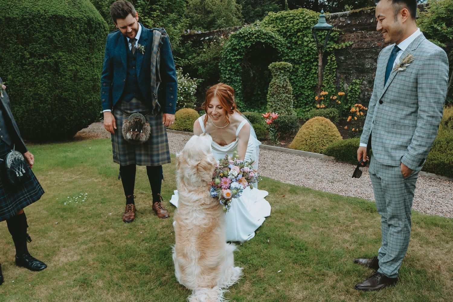 Wedding party stands on manicured garden lawn with hedges and topiary, bride wearing flowing white dress with bouquet.