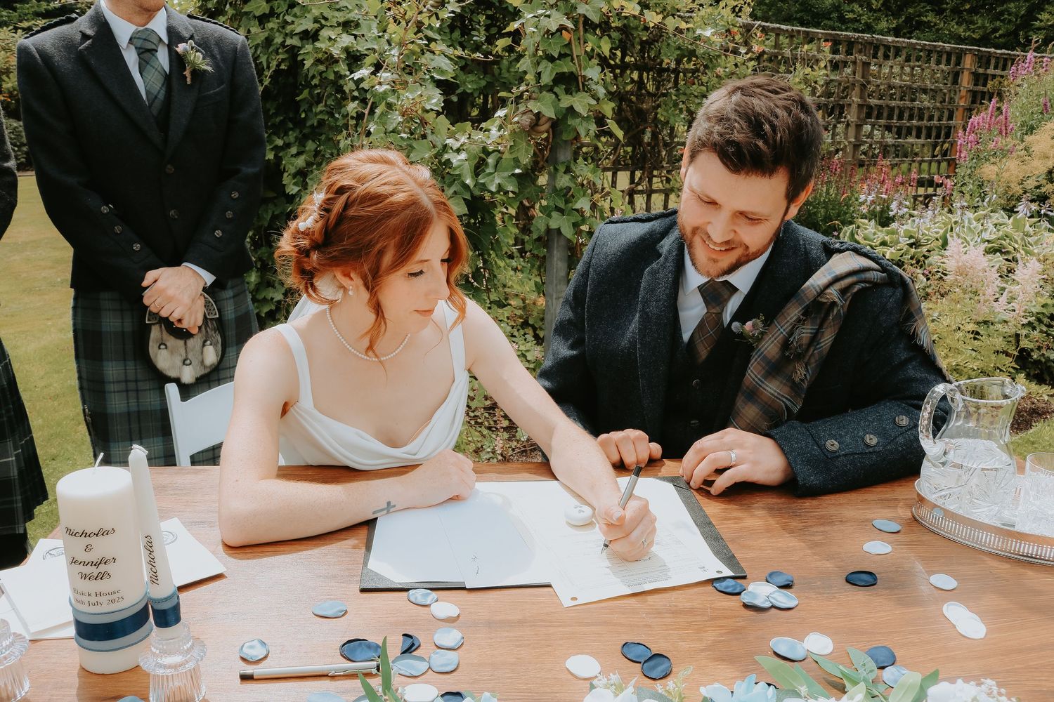Couple signing marriage documents at outdoor garden ceremony while seated at decorated table in sunlight.