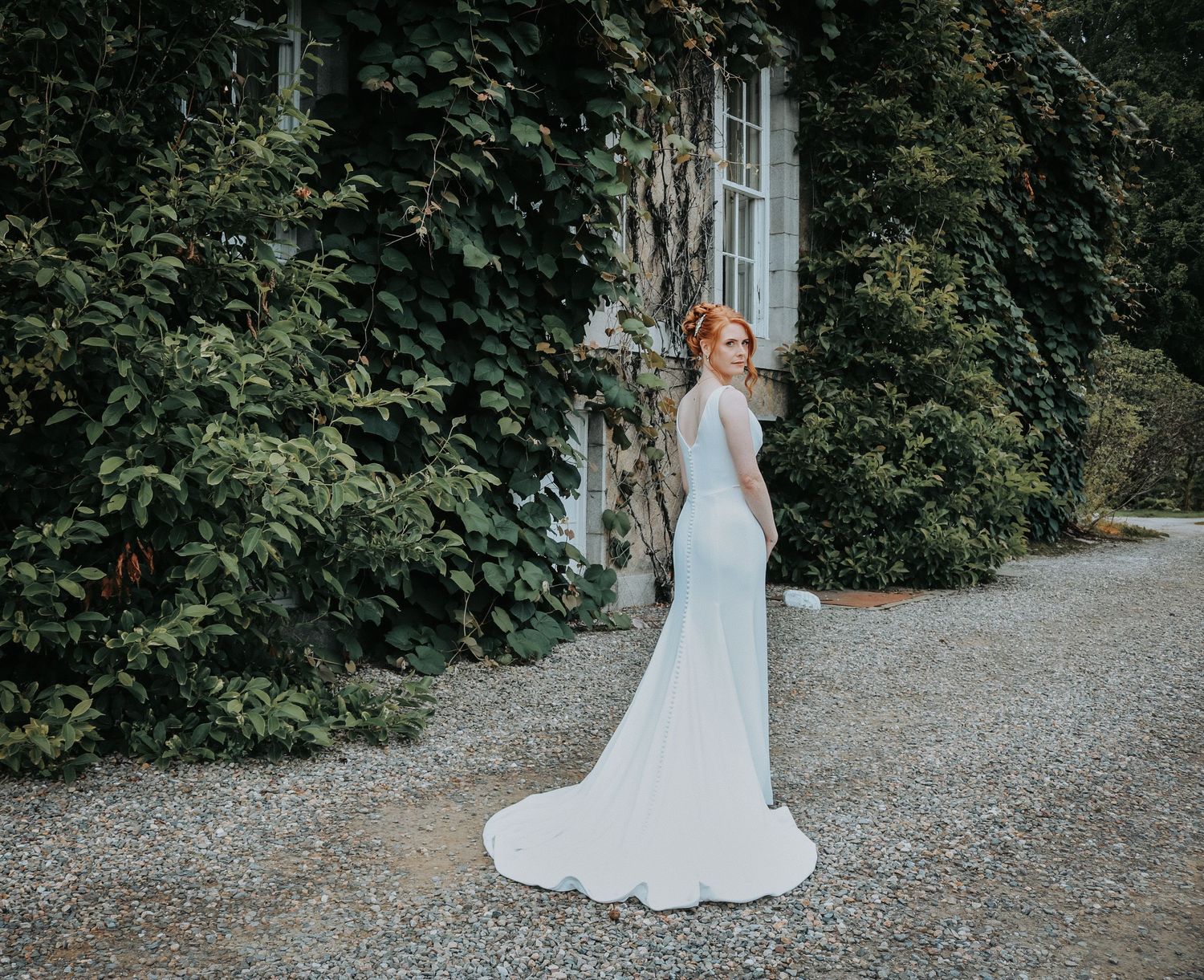 Elegant bride in flowing white gown stands on gravel path by rustic stone building covered in climbing vines.