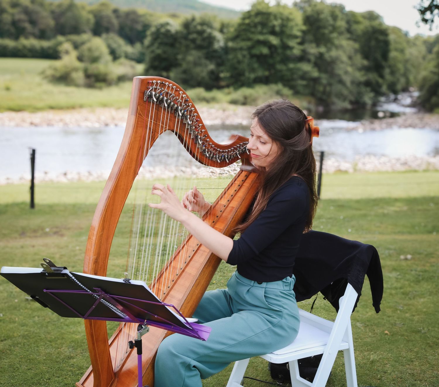 Musician plays a wooden Celtic harp outdoors on a sunny day by a lake with rolling hills in the background.
