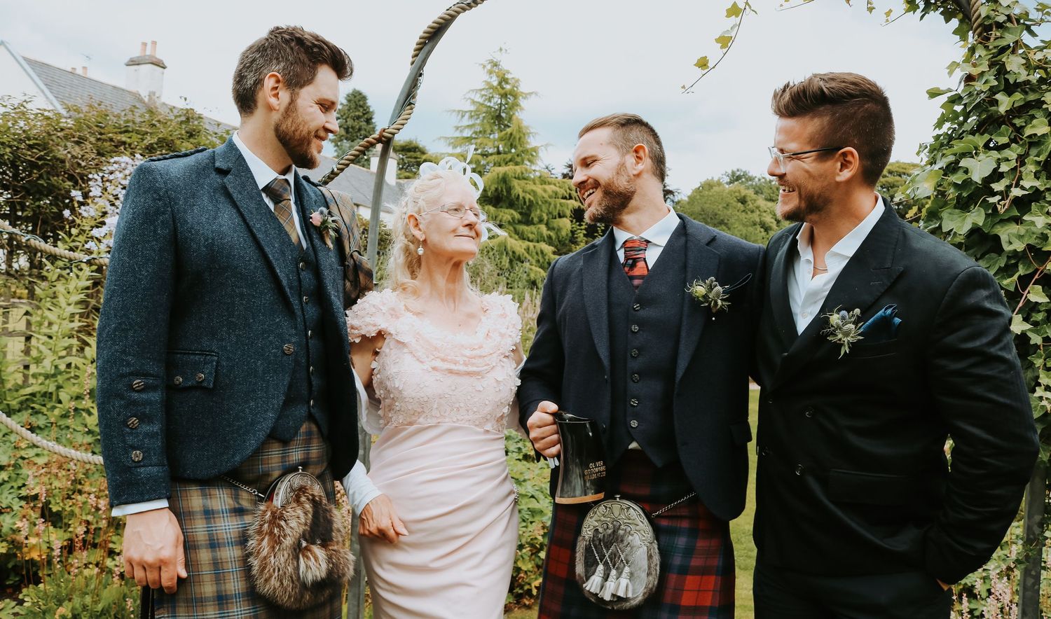 Wedding party members sharing a candid moment while wearing formal attire including traditional Scottish kilts outdoors.