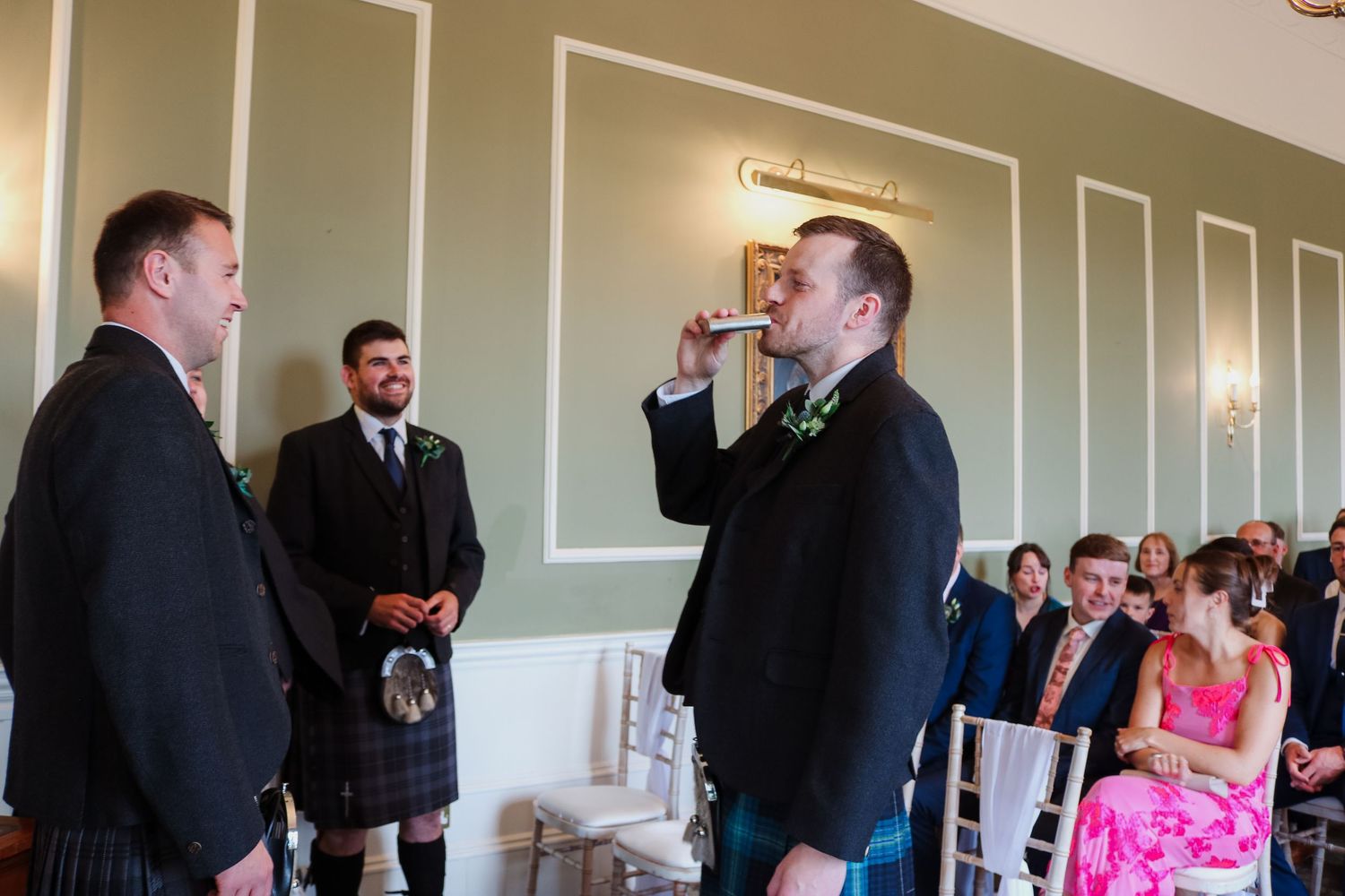 Three people standing at front of ceremony room, one in Scottish kilt attire, with seated guests visible in background.