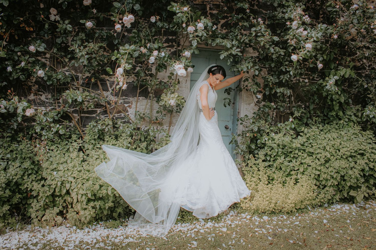 An elegant bride in a flowing white wedding gown with long veil twirls in a garden setting surrounded by greenery.