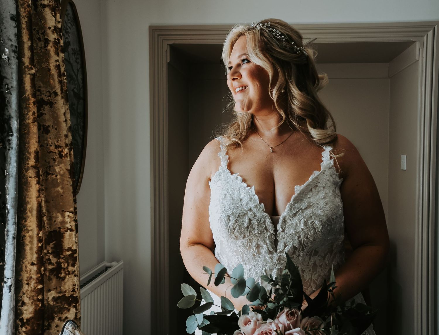 A bride wearing an elegant lace wedding gown holds a blush and eucalyptus bouquet while smiling in natural light.
