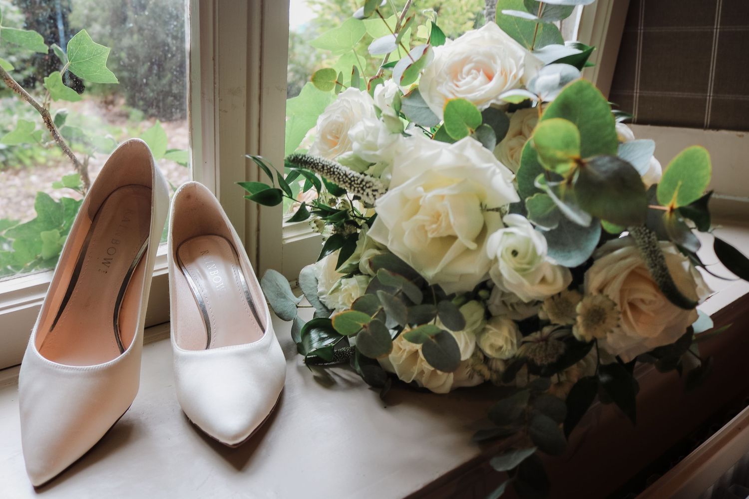 A bridal bouquet and nude wedding heels arranged with white roses and eucalyptus greenery against a light background.