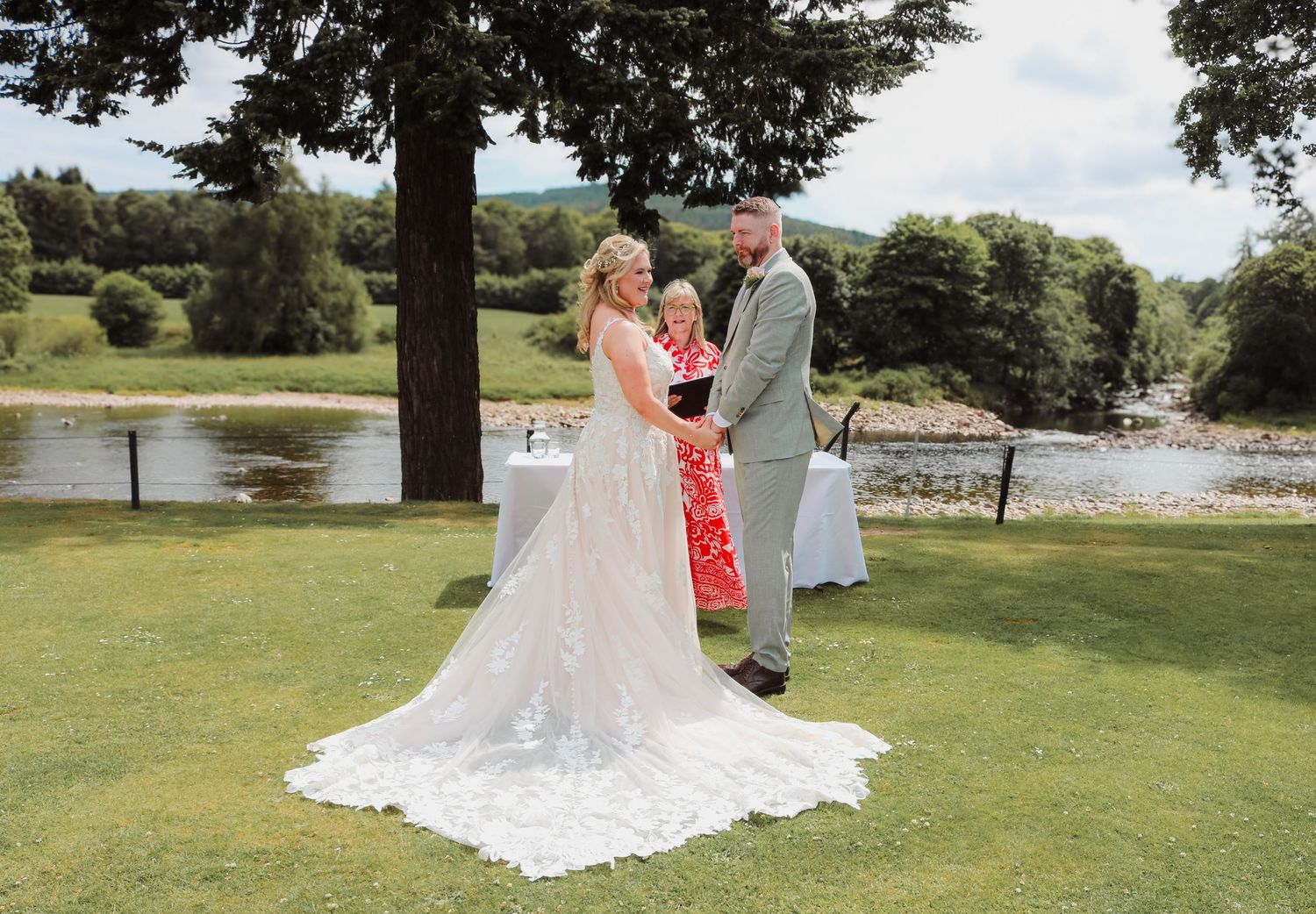 A romantic outdoor wedding ceremony takes place on a lush green lawn overlooking a serene lake on a sunny summer day.