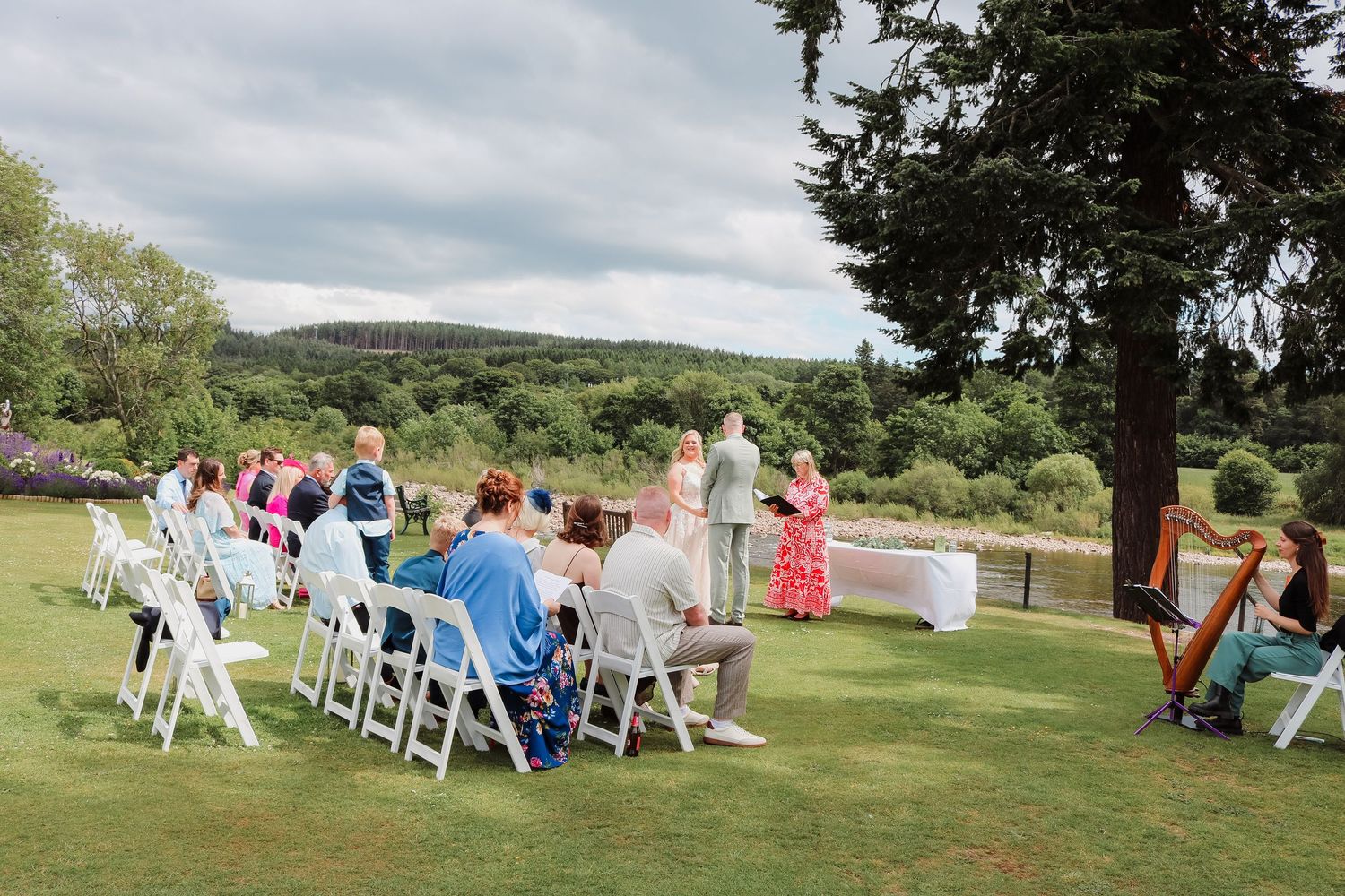Outdoor garden wedding ceremony with guests seated on white chairs and a harpist performing on a green lawn with rolling hills.