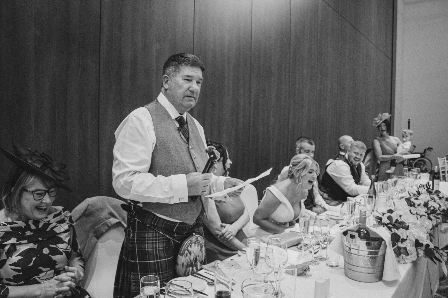 A person in traditional Scottish attire delivers a speech at a formal dinner reception with a decorated table setting.