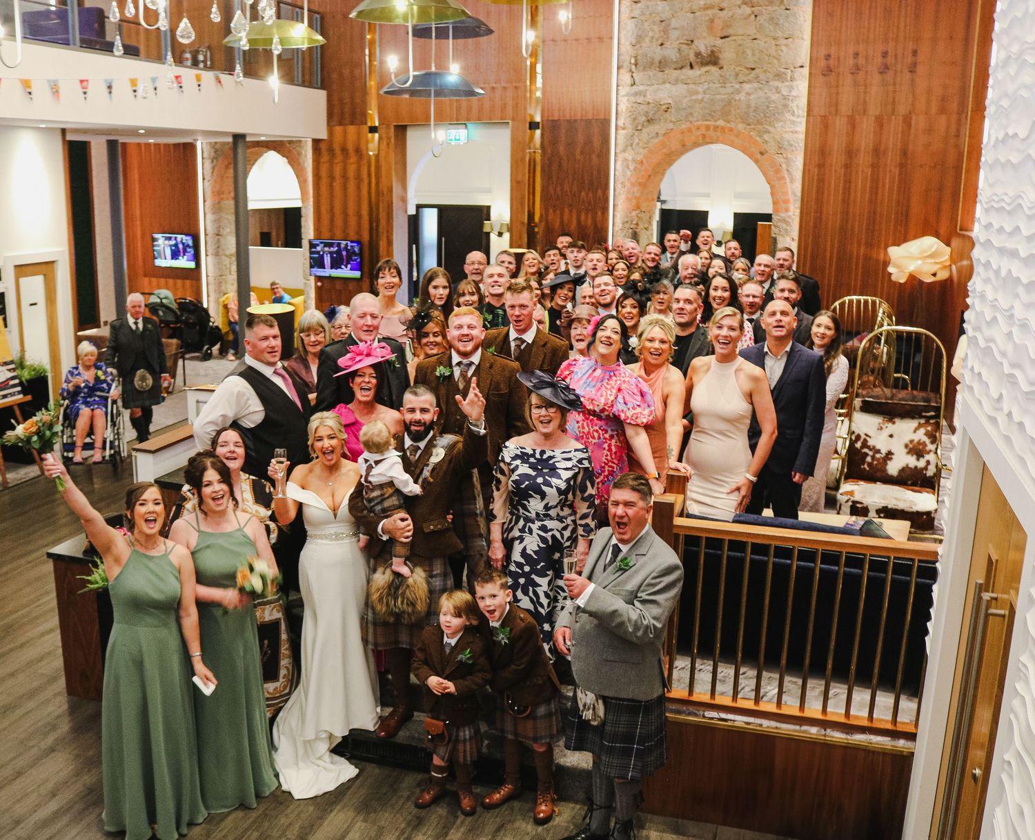 Wedding guests react with excitement and joy while looking and pointing at something off camera during a reception.