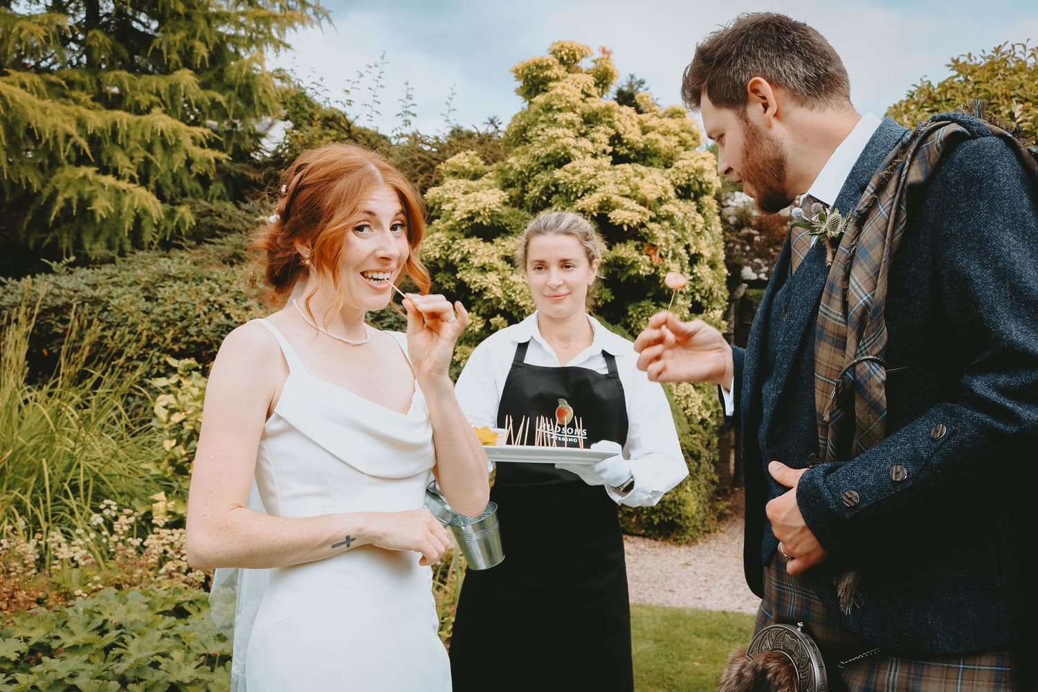 A couple taste samples from a food tray held by a server in a garden setting during a sunny outdoor event.