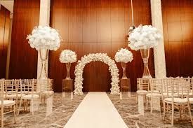 Elegant indoor wedding ceremony setup with white floral arch and tall arrangements along white carpeted aisle.