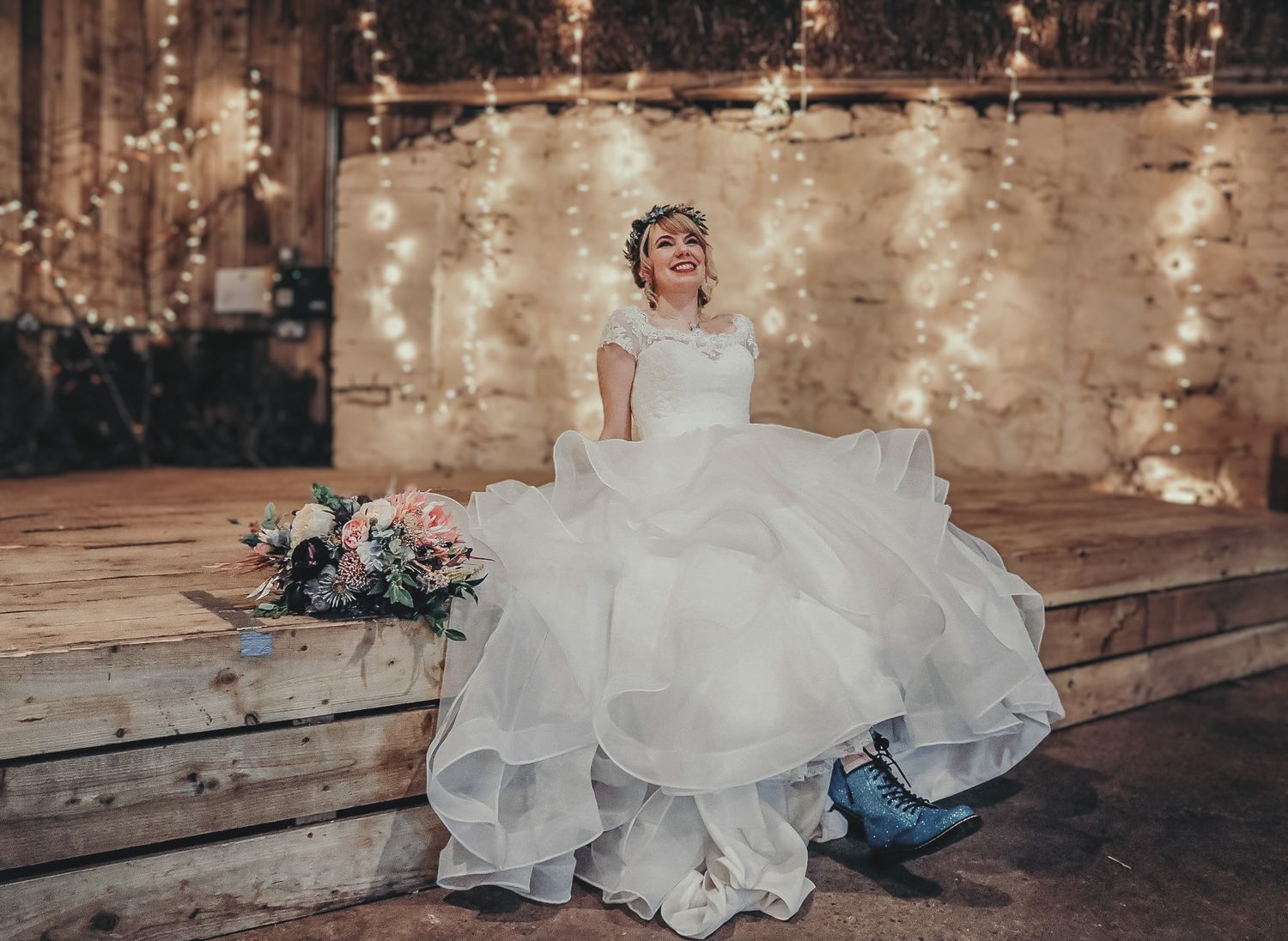A bride in a flowing white dress sits on wooden steps with string lights and a floral bouquet beside her.
