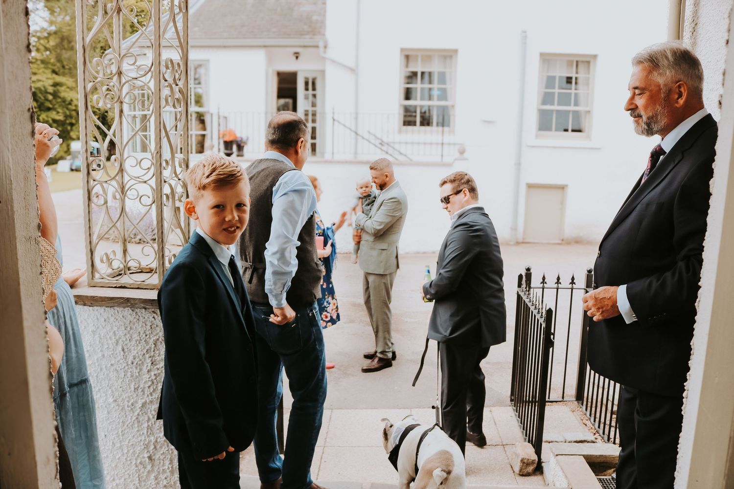 Wedding party guests stand together in black suits on a sunny porch with white columns and railings.