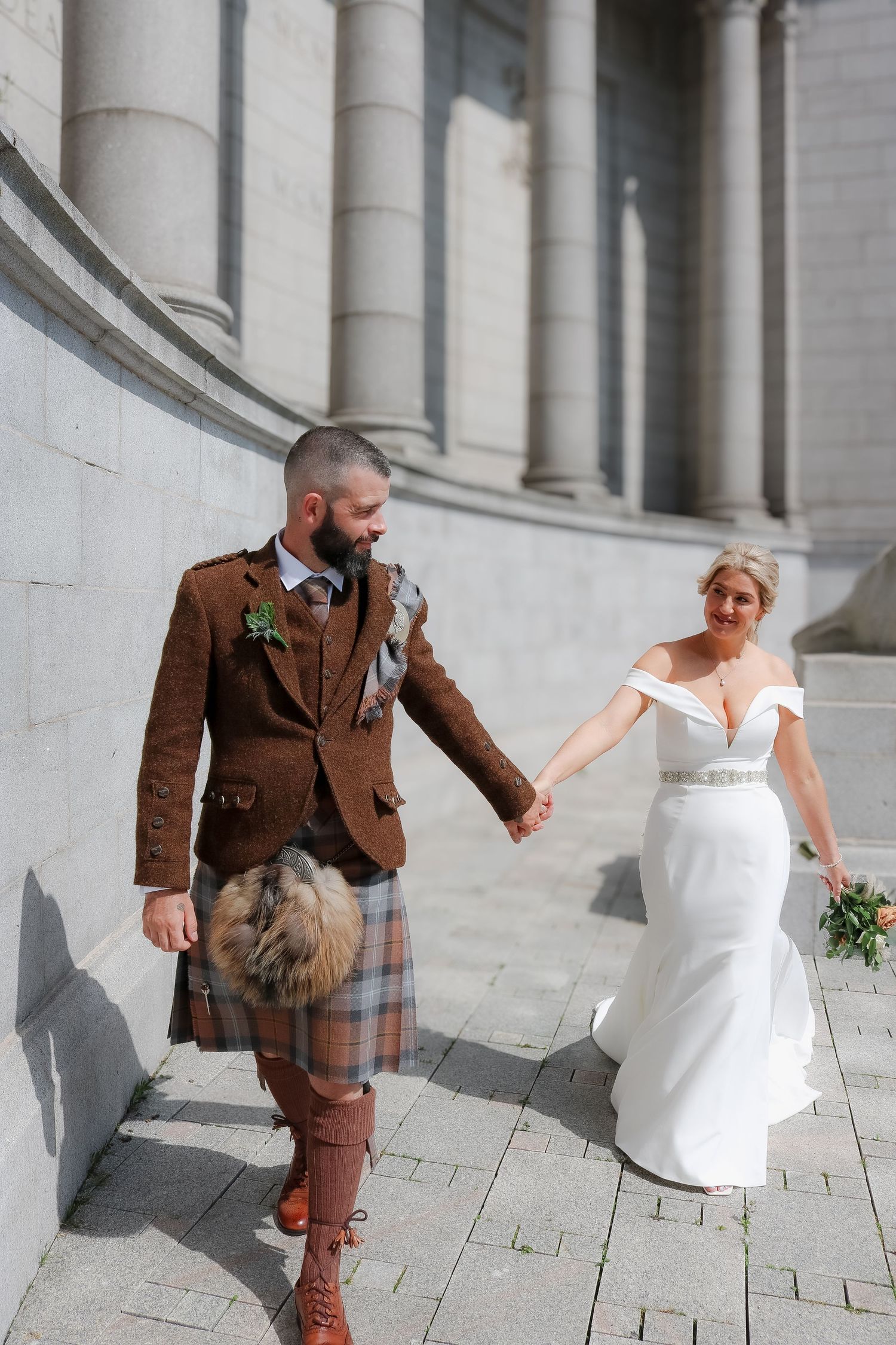 A couple in wedding attire walk together, with the groom wearing a traditional Scottish kilt and brown jacket.