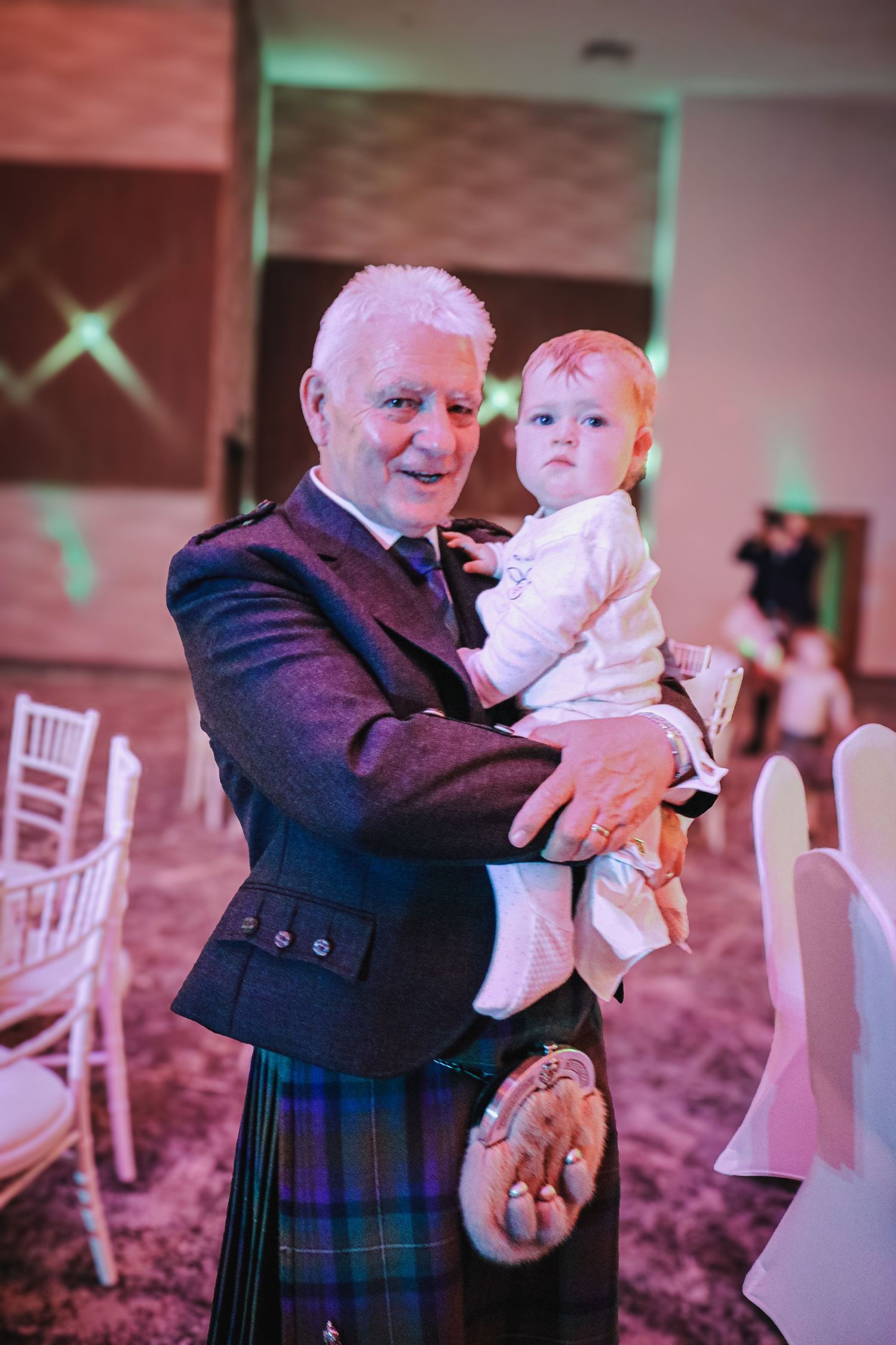 A tender moment at a wedding reception showing an older gentleman in traditional Scottish attire dancing with a small child.