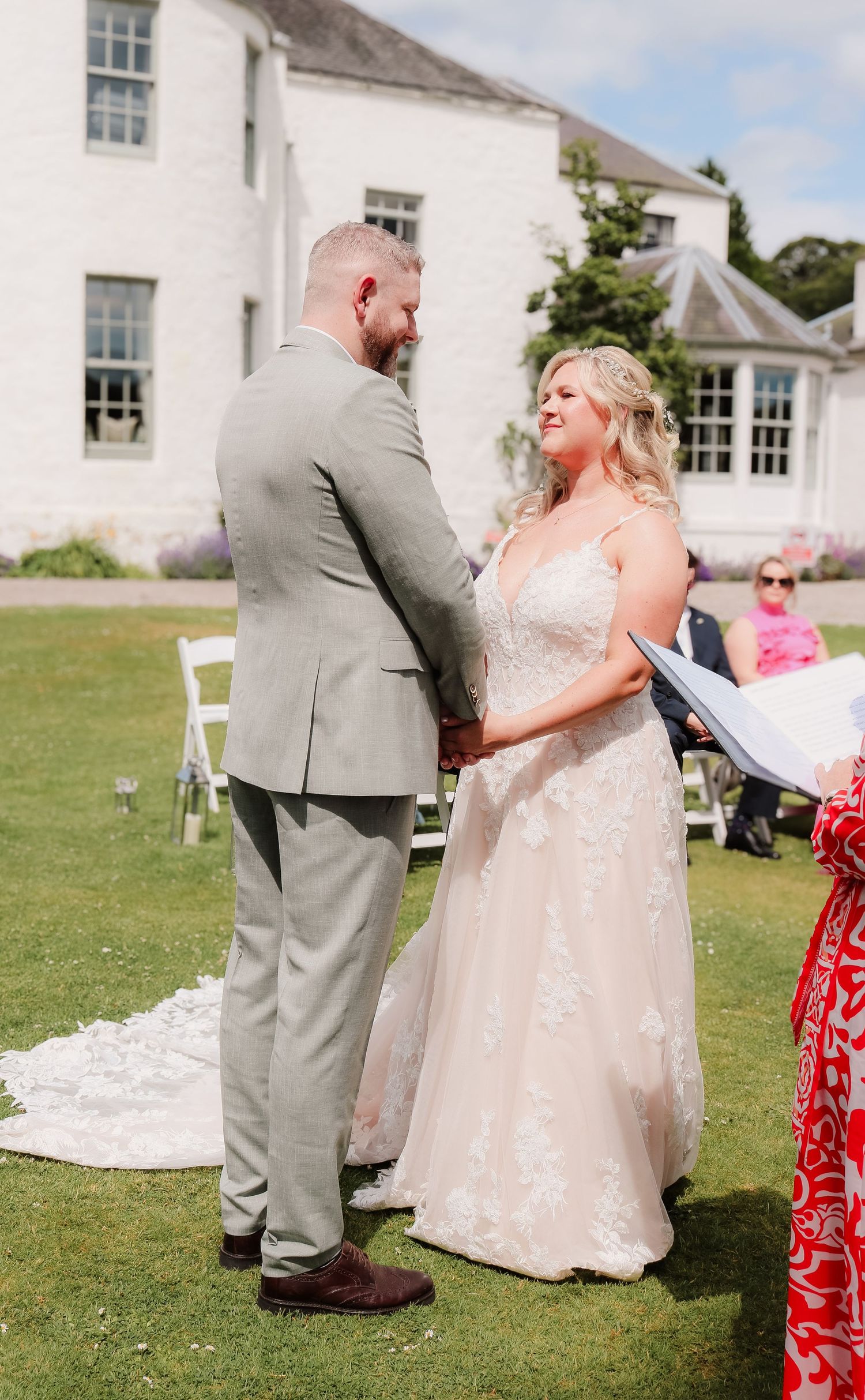 Couple exchanges vows during outdoor wedding ceremony on sunny day at white manor house with green lawn.