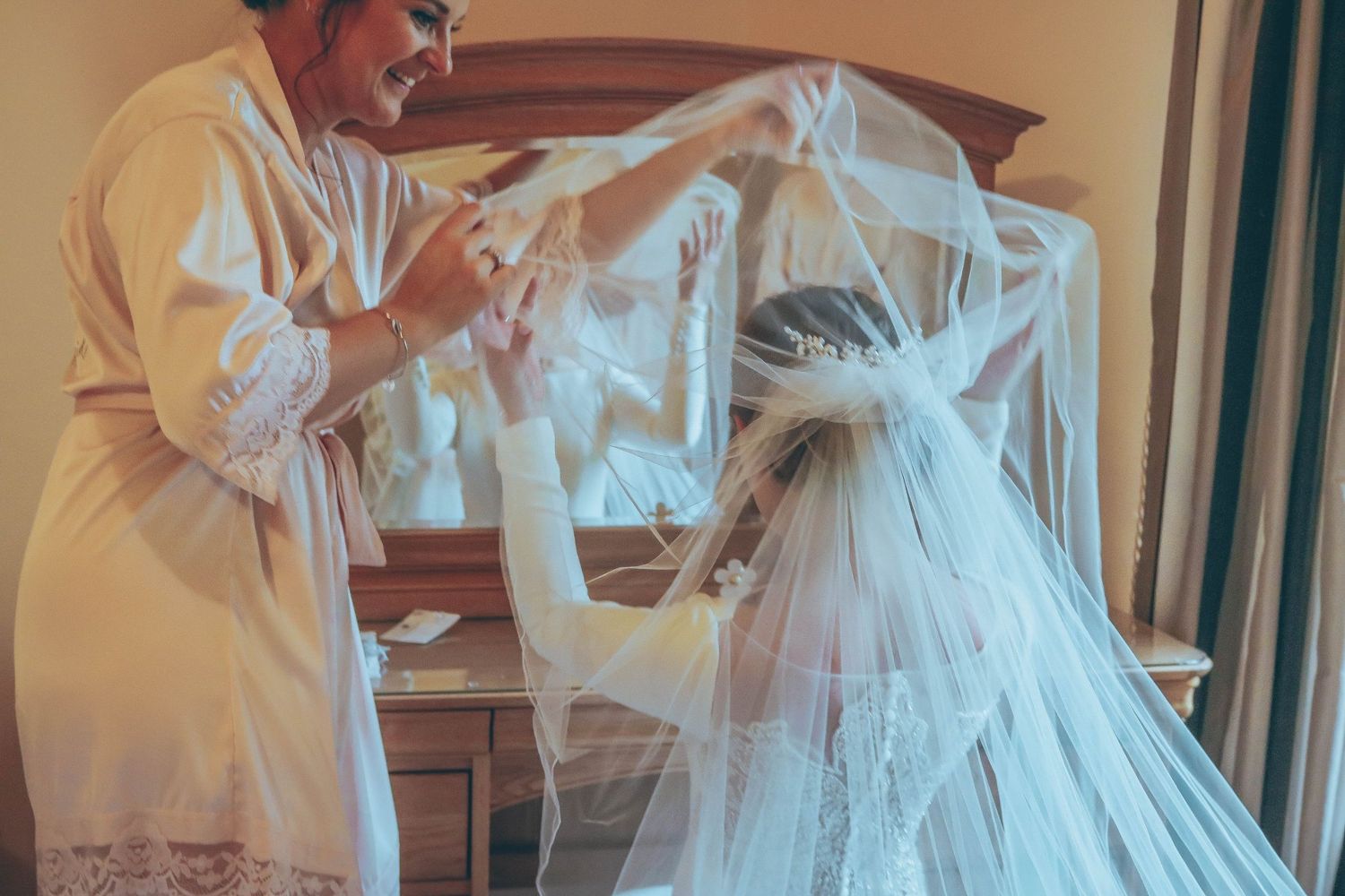 A seamstress carefully adjusts delicate white tulle and lace details on an elegant wedding gown in a sunlit room.