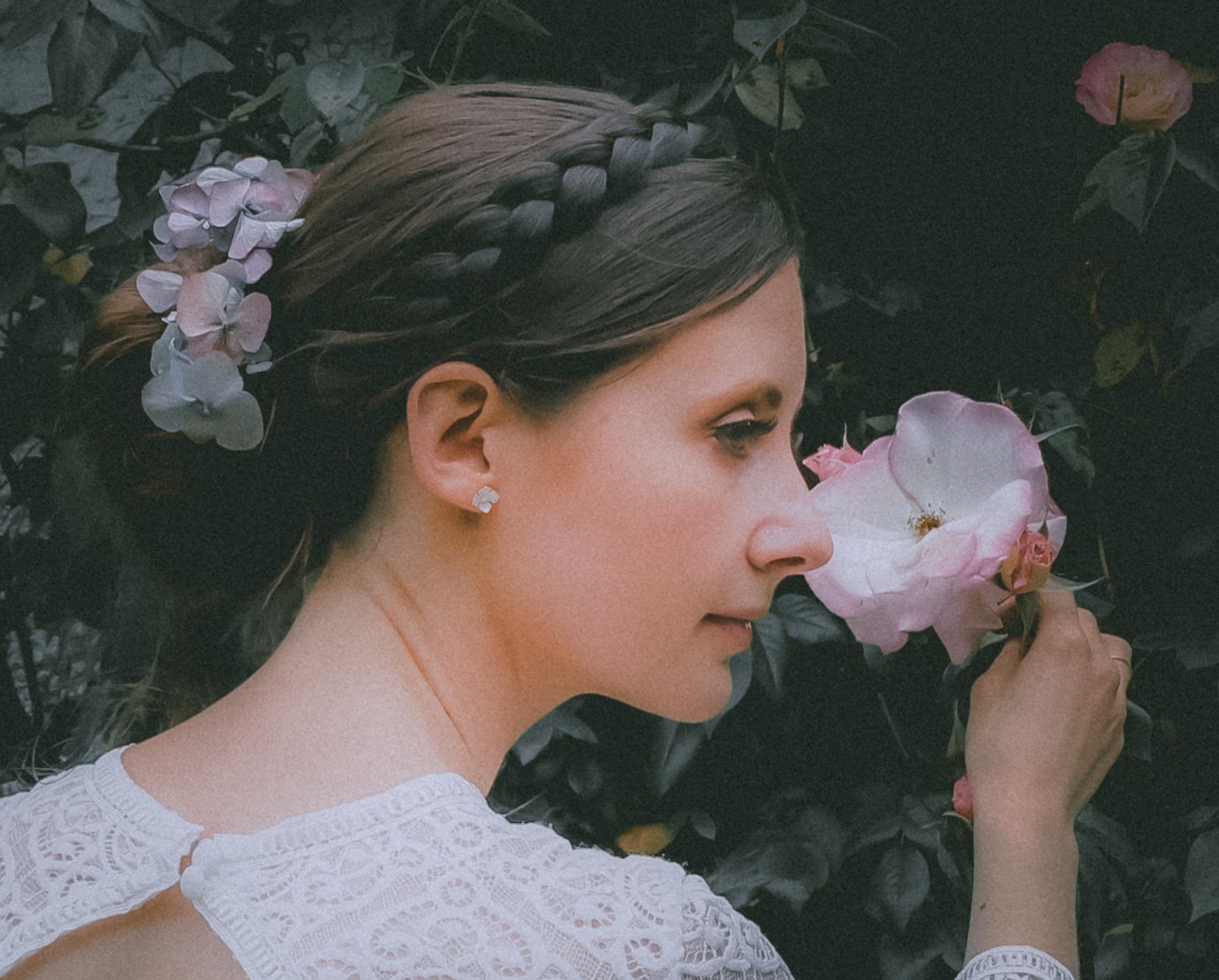 A woman in a white lace dress smells a delicate pink rose in a lush garden setting.