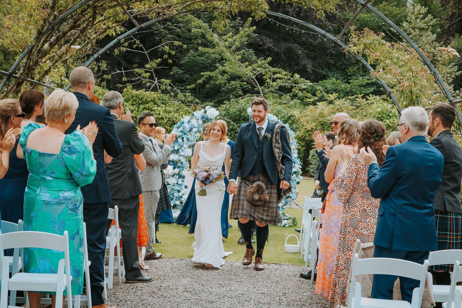 Guests mingle and celebrate during an outdoor garden wedding reception with floral decorations and white chairs.