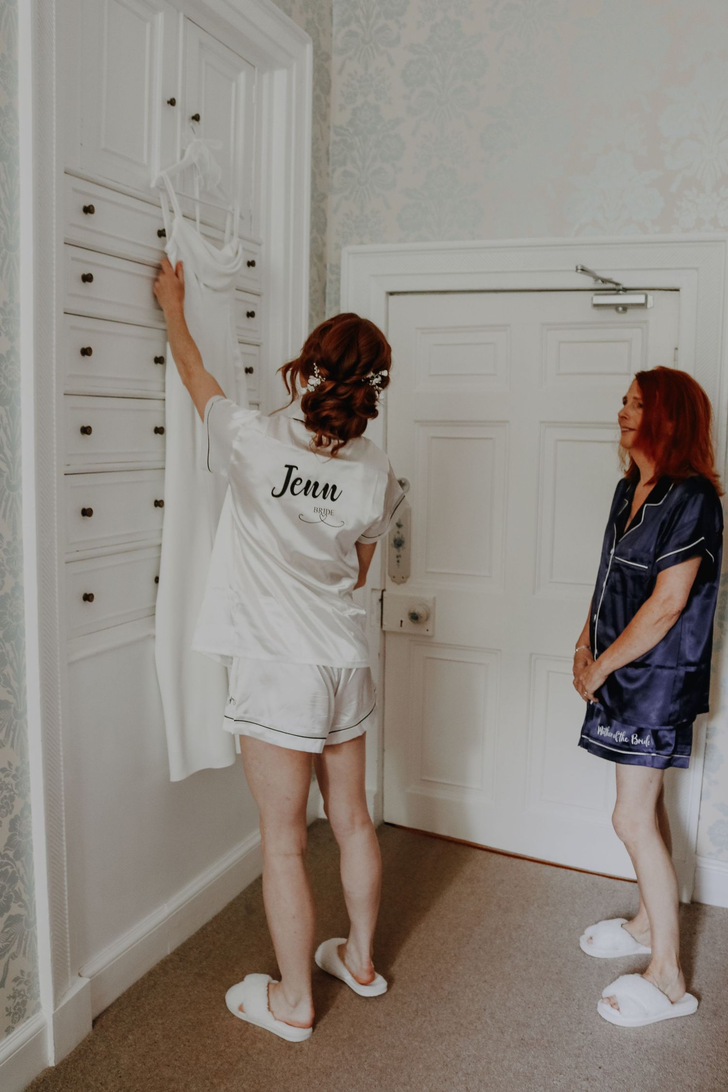 Two people in matching satin pajama sets standing in a bedroom, one reaching up to a built-in cabinet.