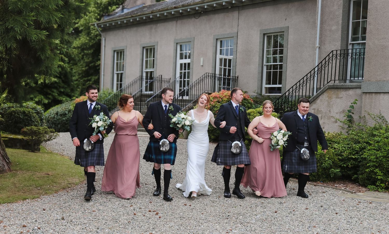 A wedding party wearing traditional Scottish kilts and formal attire poses together on a gravel path outside a building.