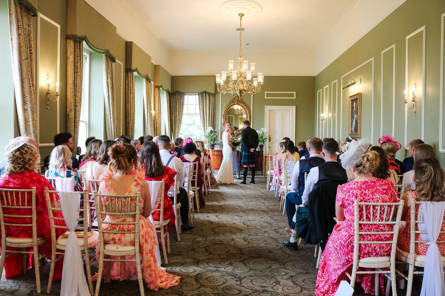 Elegant wedding ceremony in a green-walled room with chandelier lighting and rows of pink chairs for guests.
