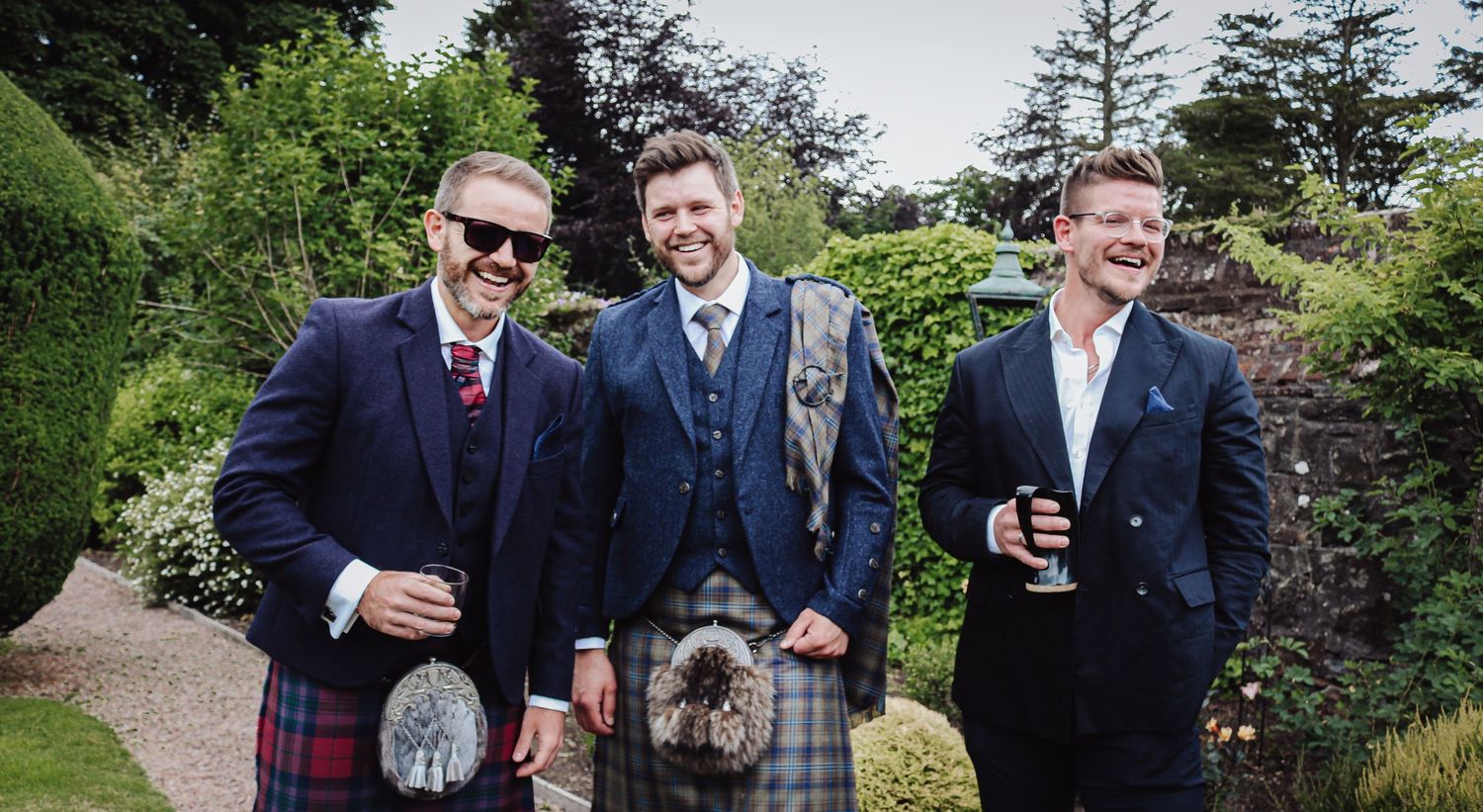 A group of formally dressed guests mingle and laugh at an outdoor social gathering in a garden setting.