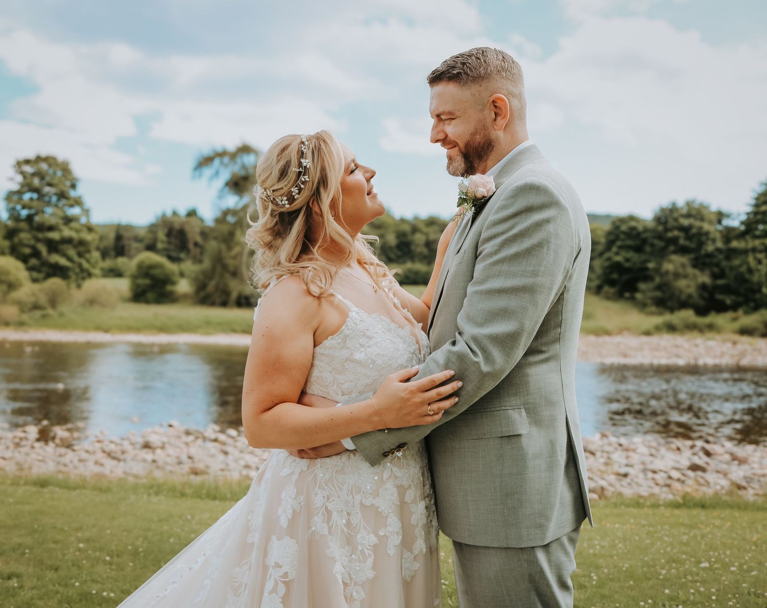 A couple in wedding attire embrace lovingly by a scenic lake on their special day, surrounded by lush greenery.