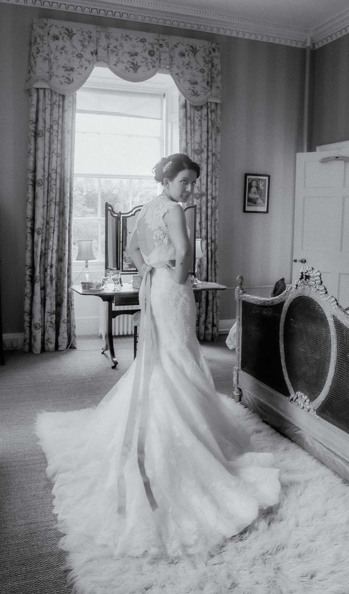 A bride in an elegant mermaid-style wedding gown poses in front of a window in an ornate vintage room.