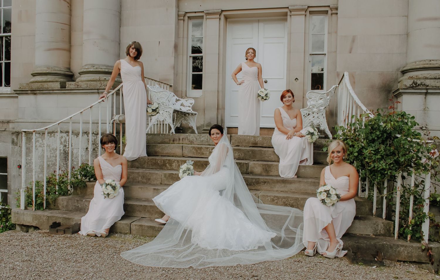 An elegant bridal party in white dresses poses gracefully on the grand steps of a historic mansion.