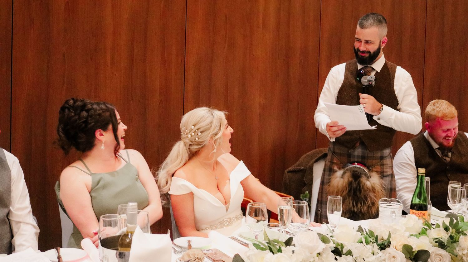 Wedding reception scene with guests seated at a decorated table while a server takes orders at a banquet venue.