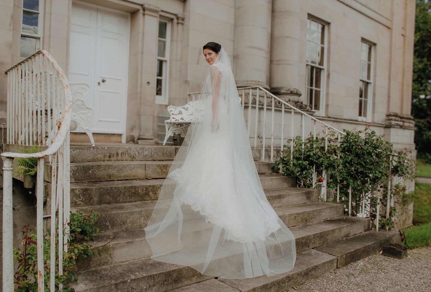 A bride in an elegant white tulle wedding gown stands gracefully on stone steps of a historic white building.