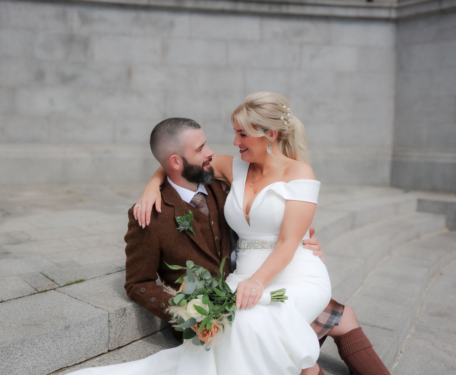 Happy newlywed couple in white dress and brown suit share a playful moment while sitting on stone steps.