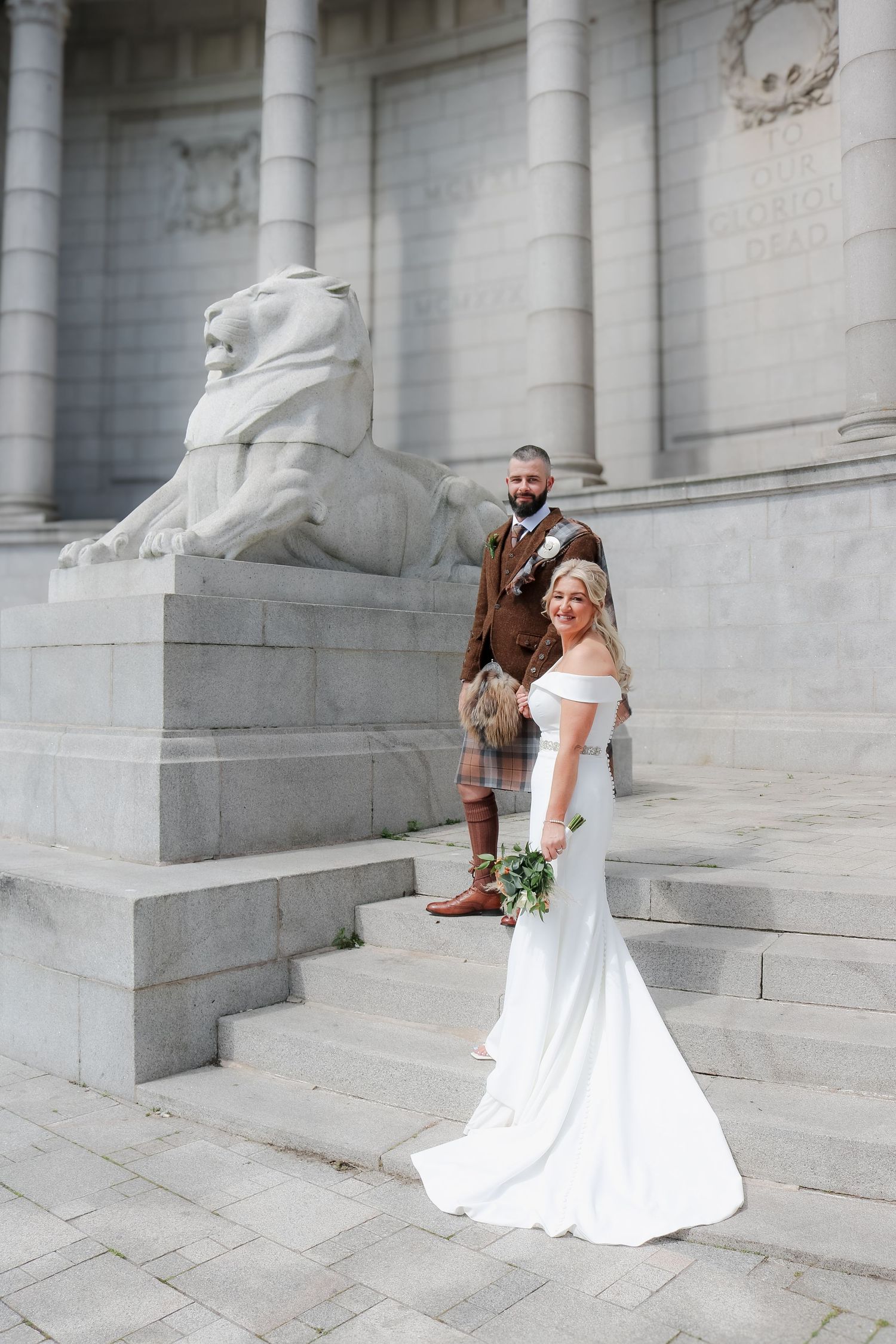 Elegant wedding couple poses on stone steps beside a majestic lion statue at a grand architectural building.