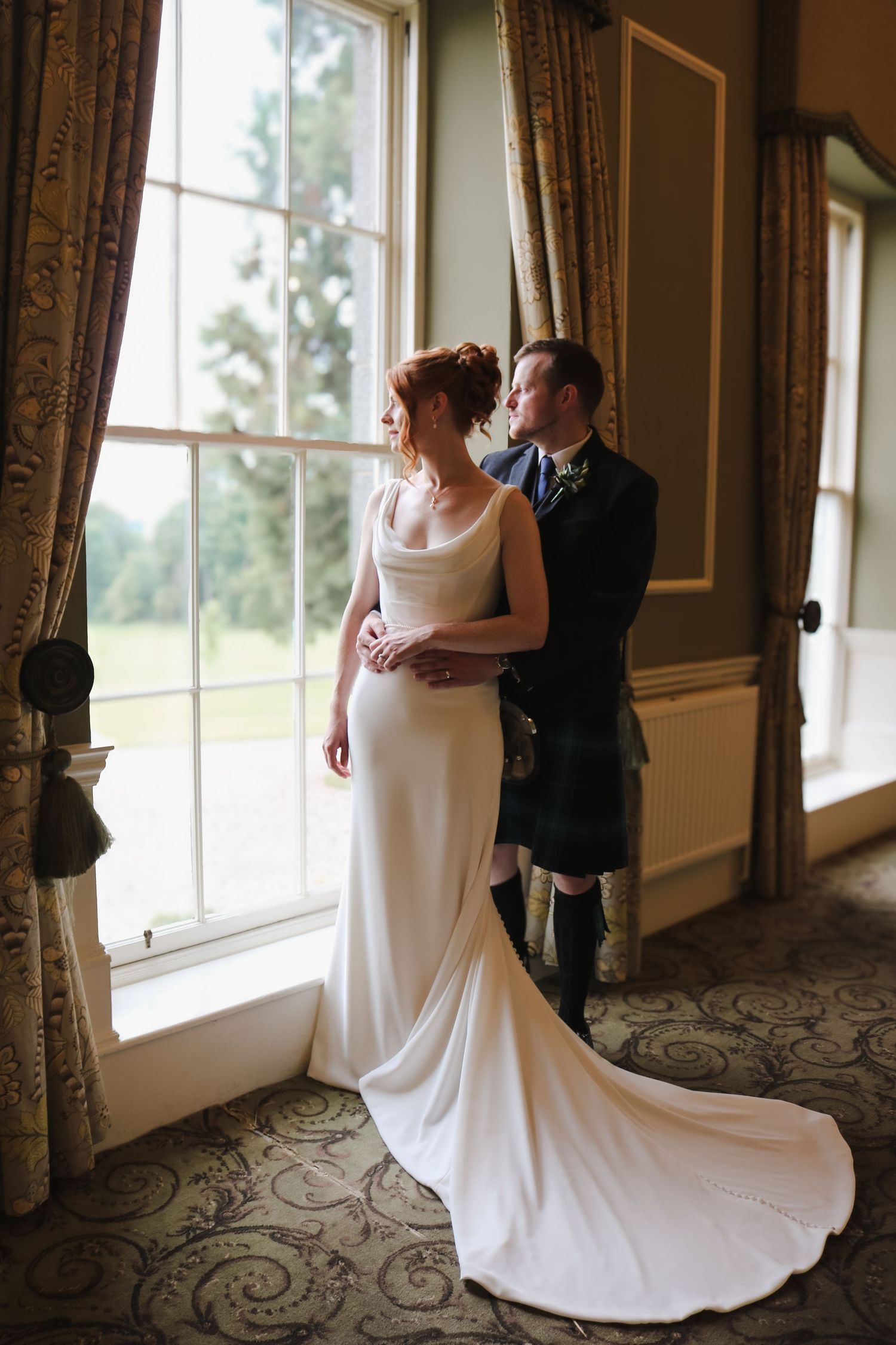 A couple shares an intimate moment by large windows in an elegant room, with the bride in a flowing white gown and groom in kilt.